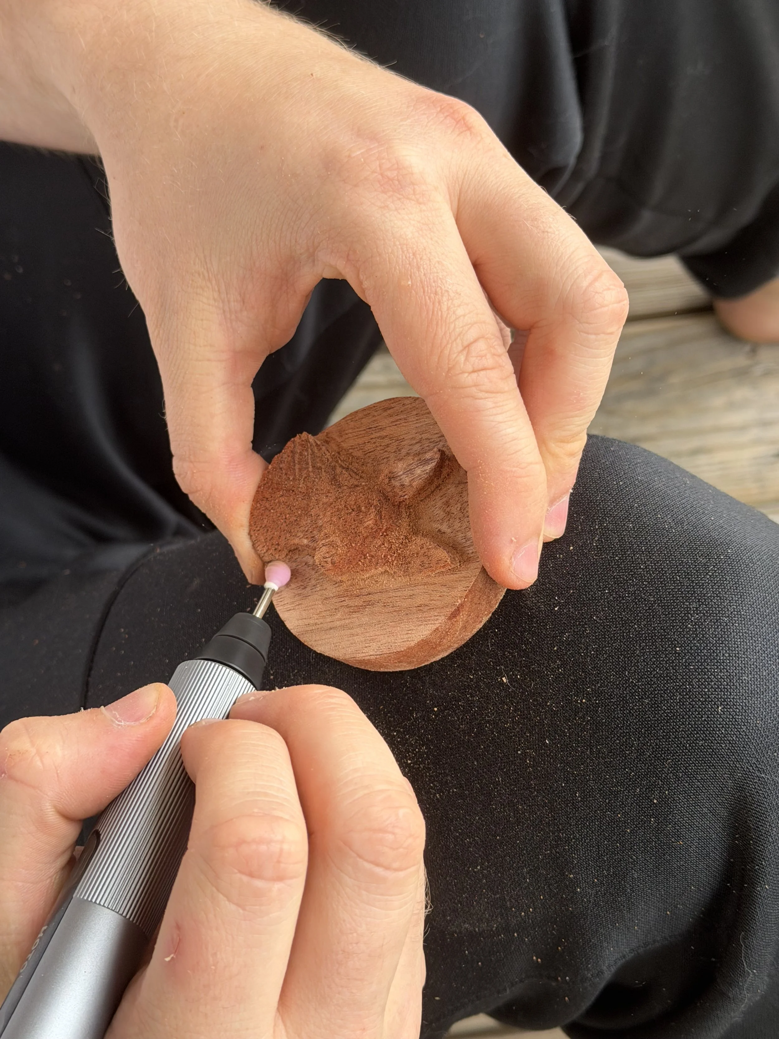 A person is carving a detailed design into a wooden object using a small rotary tool, holding the wood steady with one hand and guiding the tool with the other.