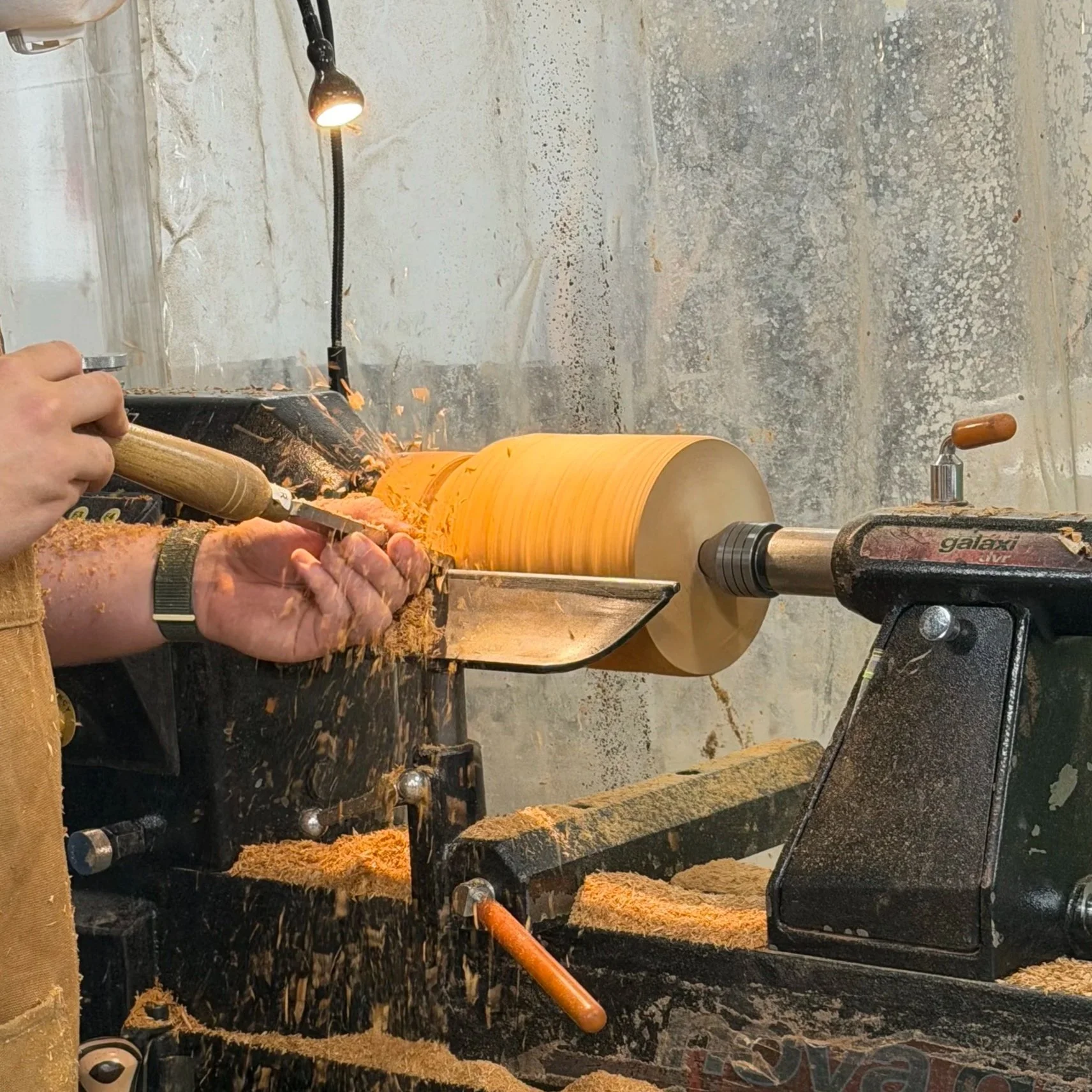 Woodturning process in a workshop, showing a person shaping a cylindrical wooden piece on a lathe with wood shavings scattered around.