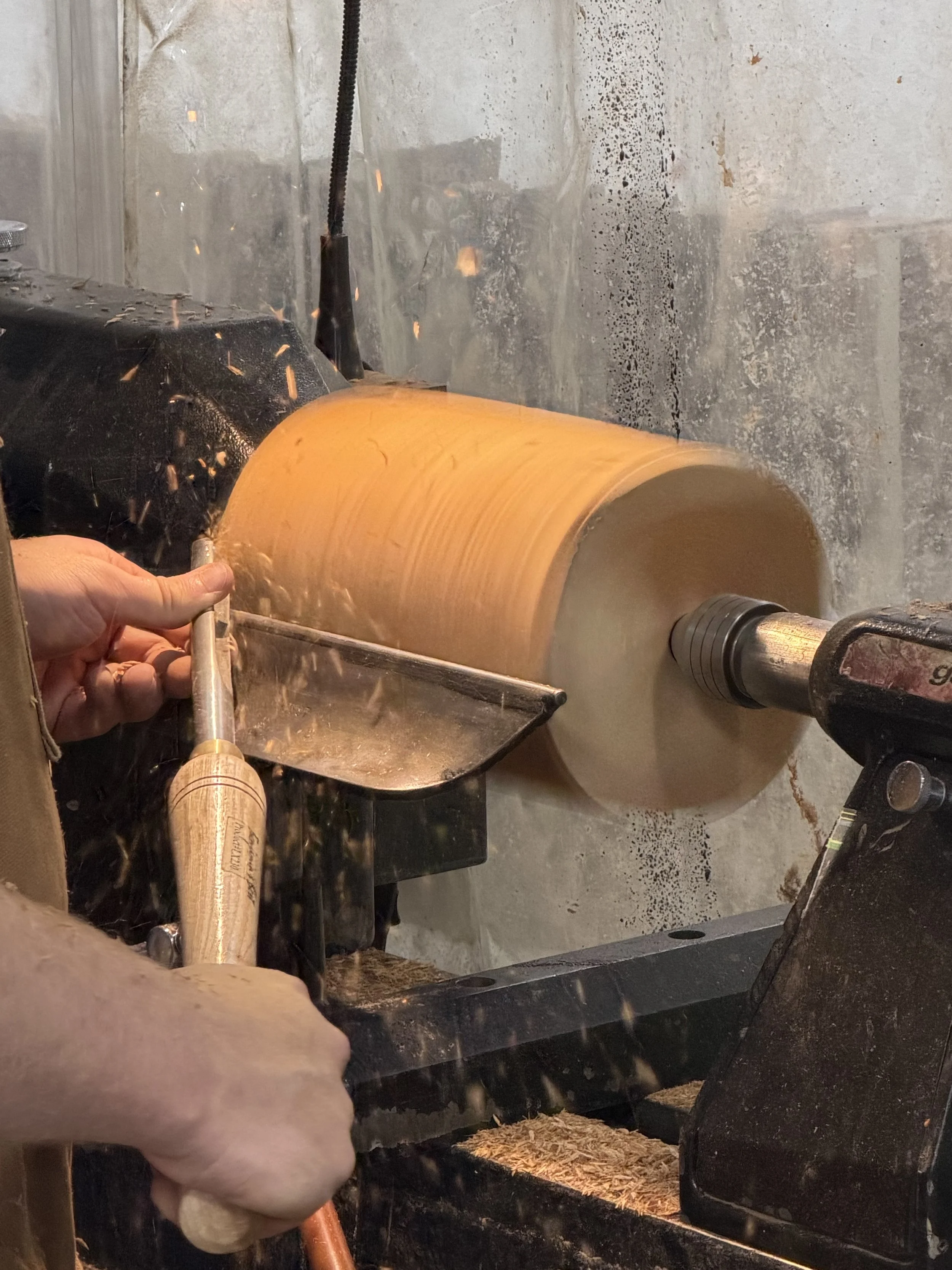 Person using a drill to cut or shape a wooden log on a workbench, with wood shavings flying around, and a transparent plastic sheet in the background.