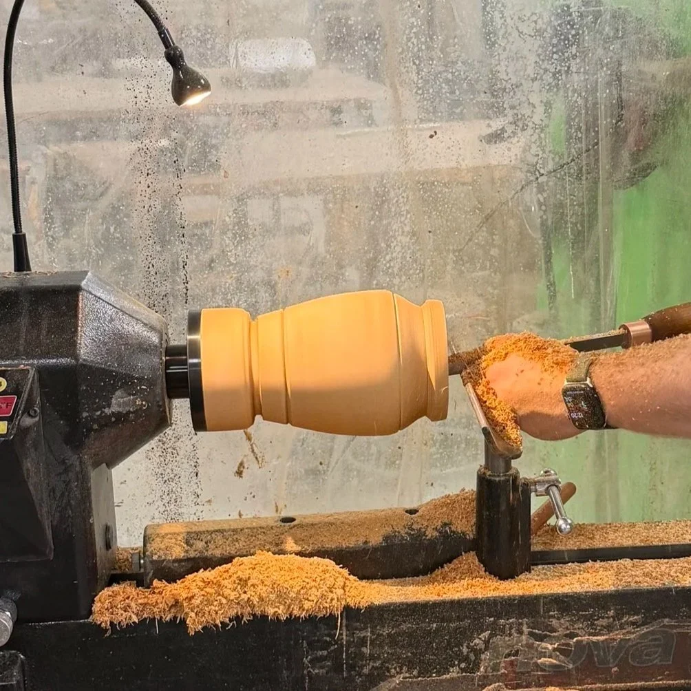 A person using a lathe to shape a wooden object, with wood shavings and dust around.