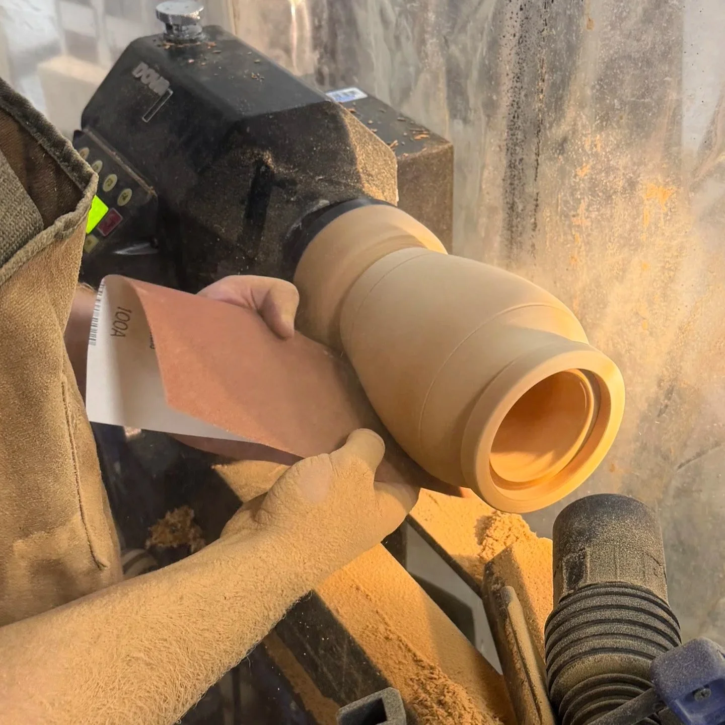 A person in a tan work glove holding a block of sandpaper and sanding a tan-colored ceramic piece on a rotary grinder in a workshop.