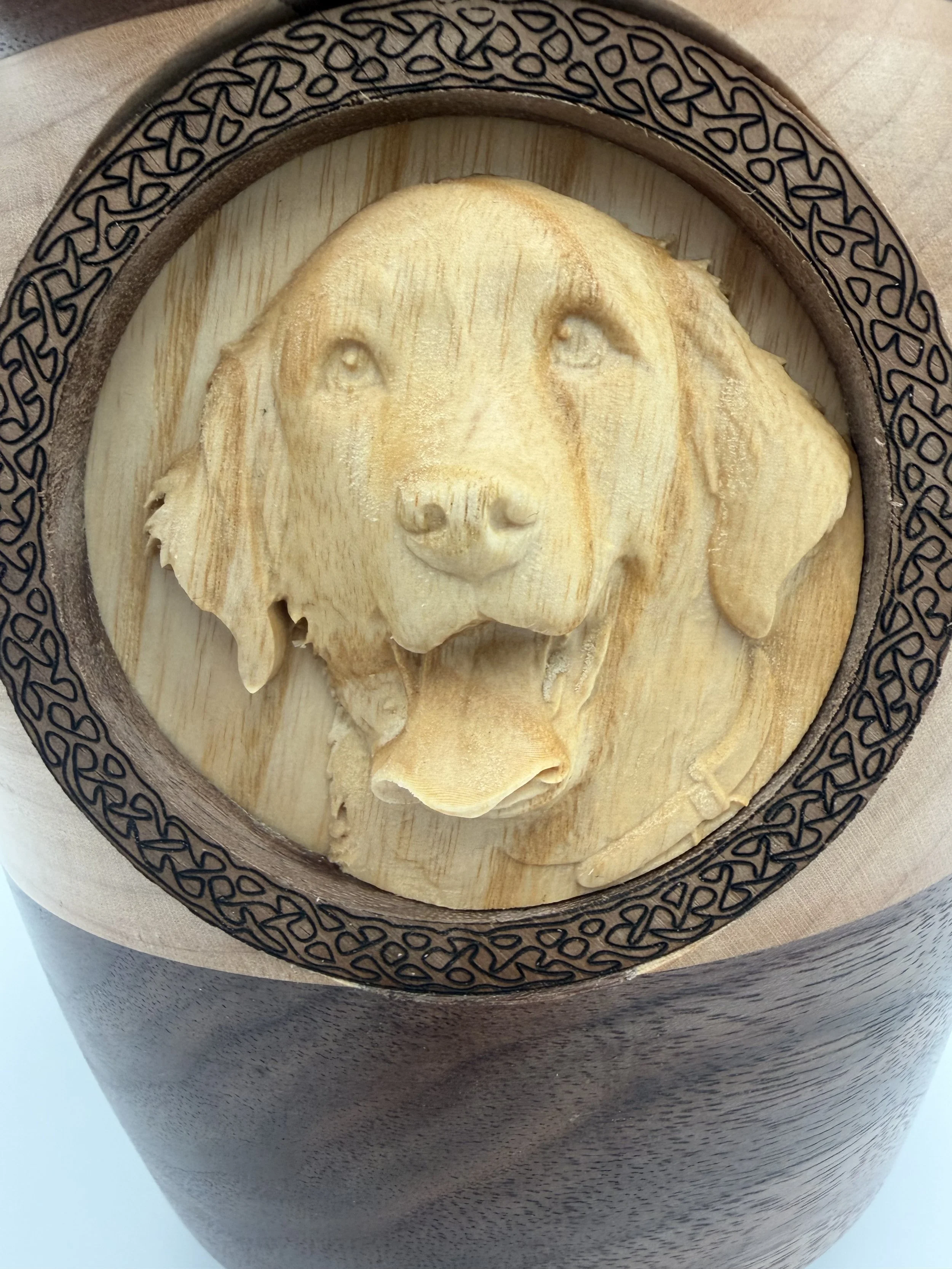 A wood carving of a dog's face on a round wooden board, placed on top of a dark rounded surface with a decorative pattern along the edge.