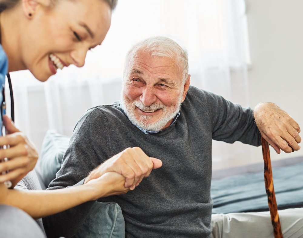 An elderly man with a white beard and gray hair sitting on a sofa, smiling at a young woman. The elderly man is holding a wooden cane with his right hand. The young woman, partially visible, is smiling and appears to be helping or supporting him, suggesting a caring interaction.