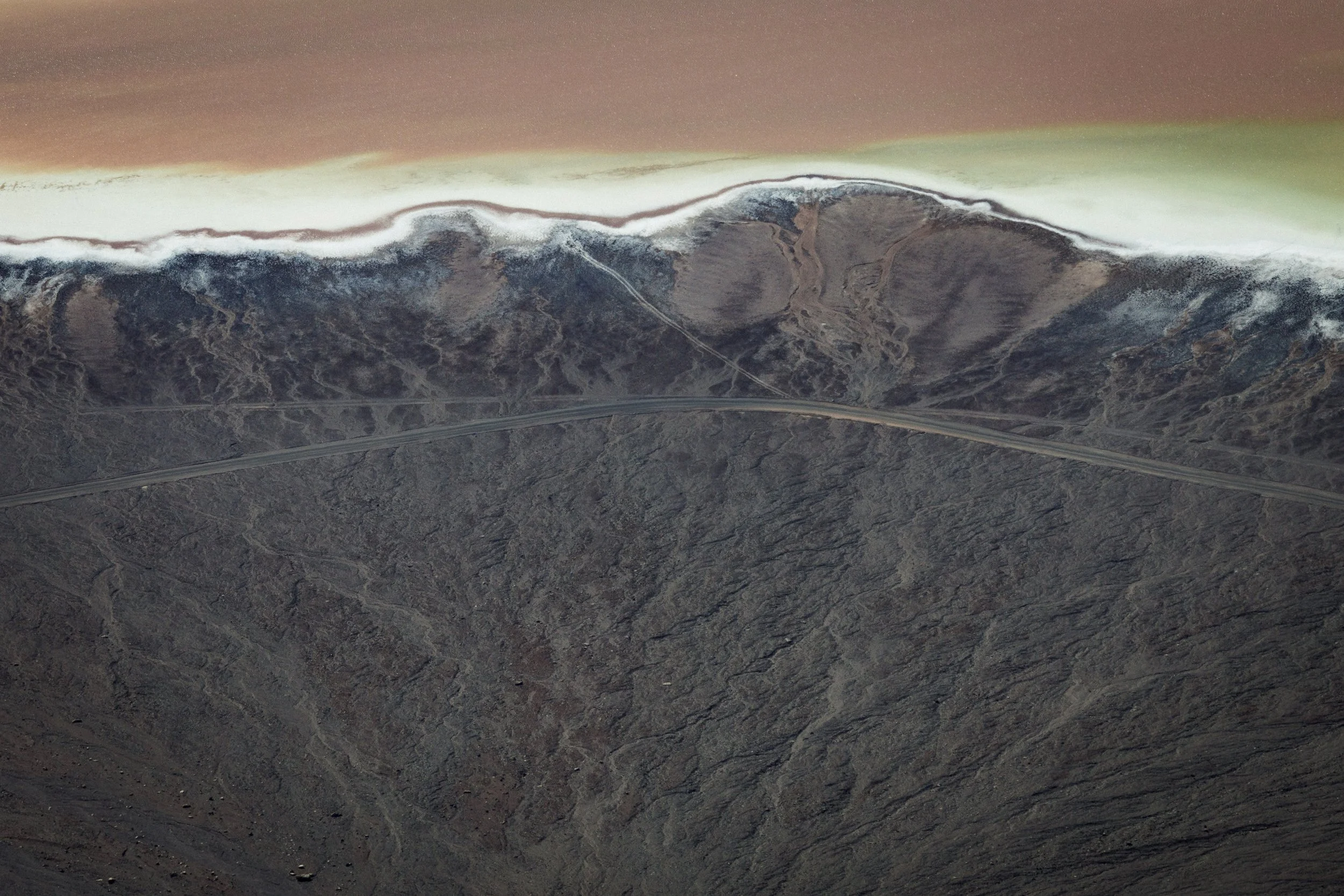 A barren mountainous landscape with dry, rocky terrain and winding dirt roads beneath a colorful, dusty sky with a gradient of brown, green, and light hues.