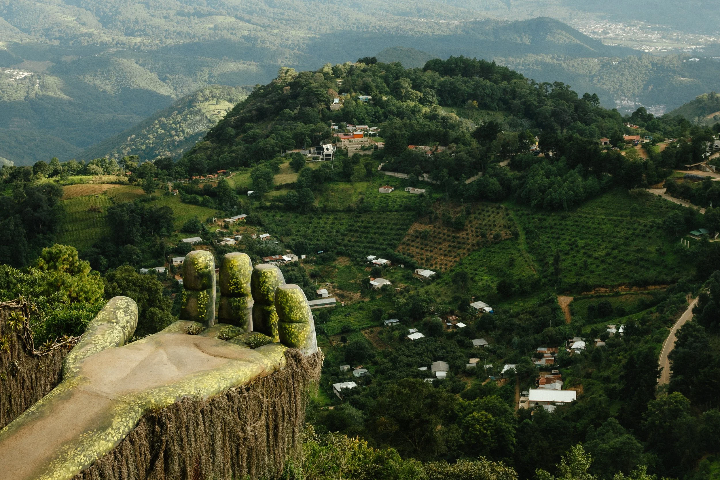 A scenic view of green hills and a small village, taken from a high vantage point, with large moss-covered hand sculptures in the foreground.