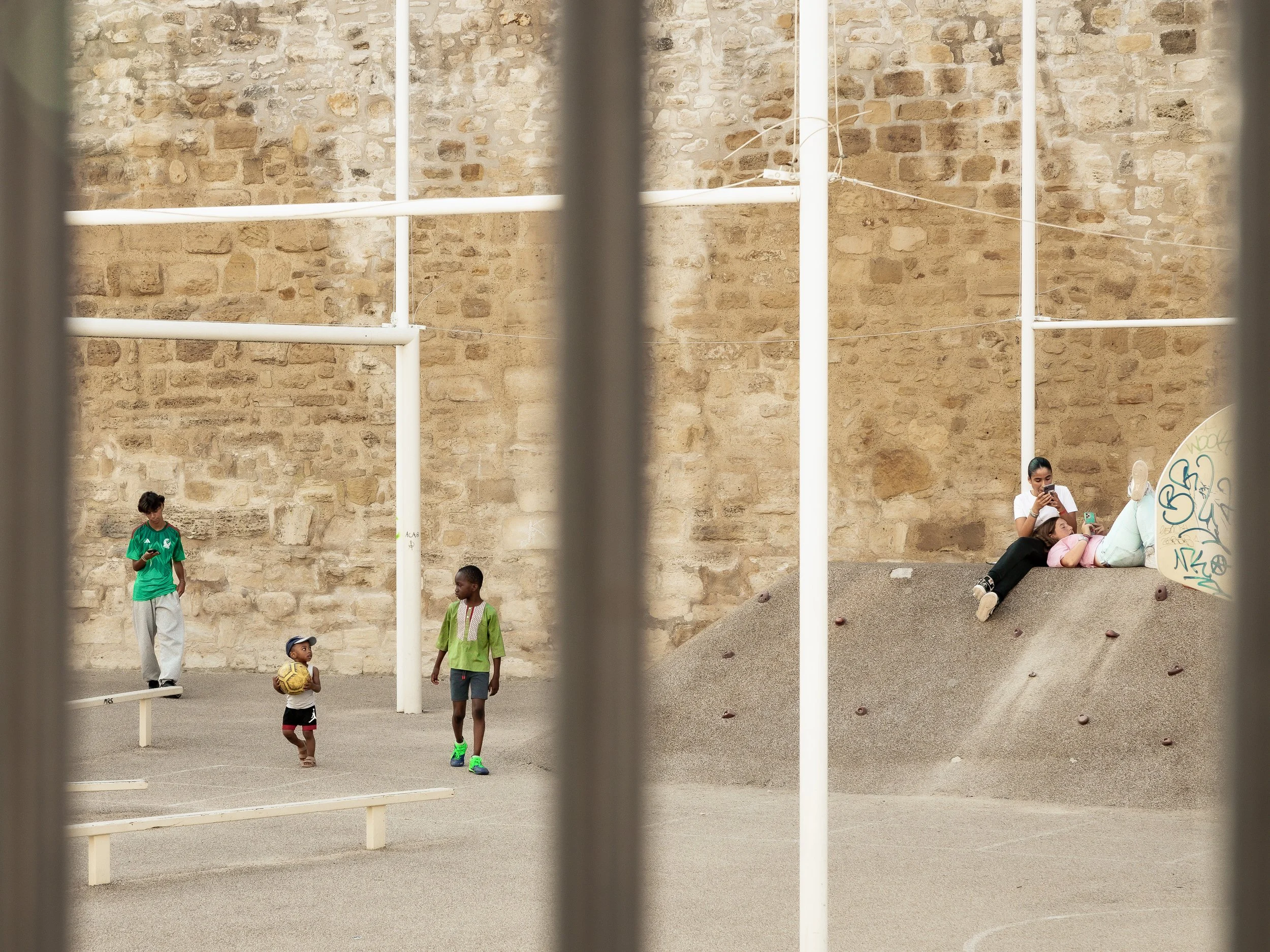 Children playing in an urban playground with a stone wall background and some adults sitting on a small wall. The photo is taken through metal bars.