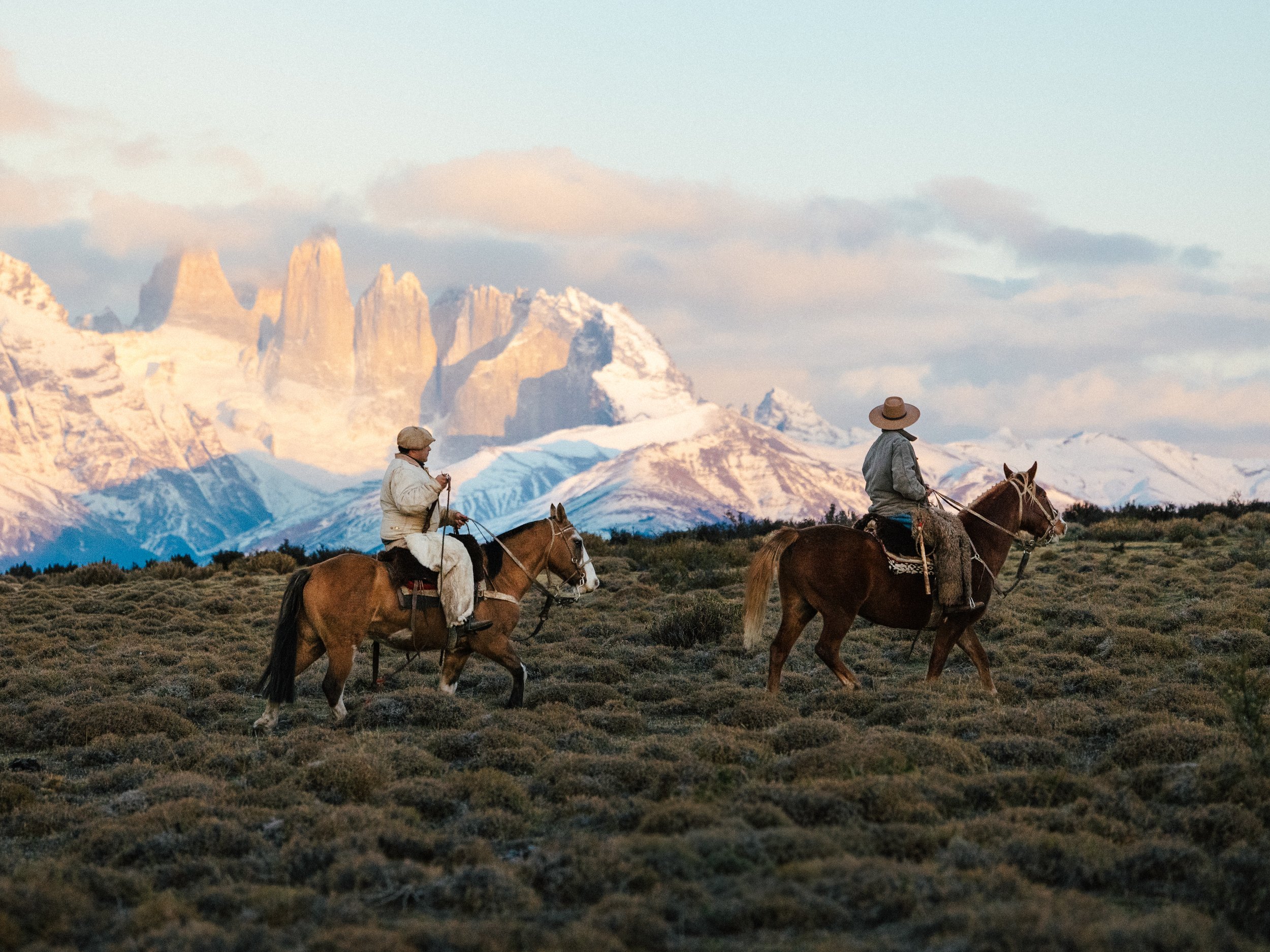 Two people riding horses in a flat, shrub-covered landscape with snow-capped mountains and a partly cloudy sky in the background.