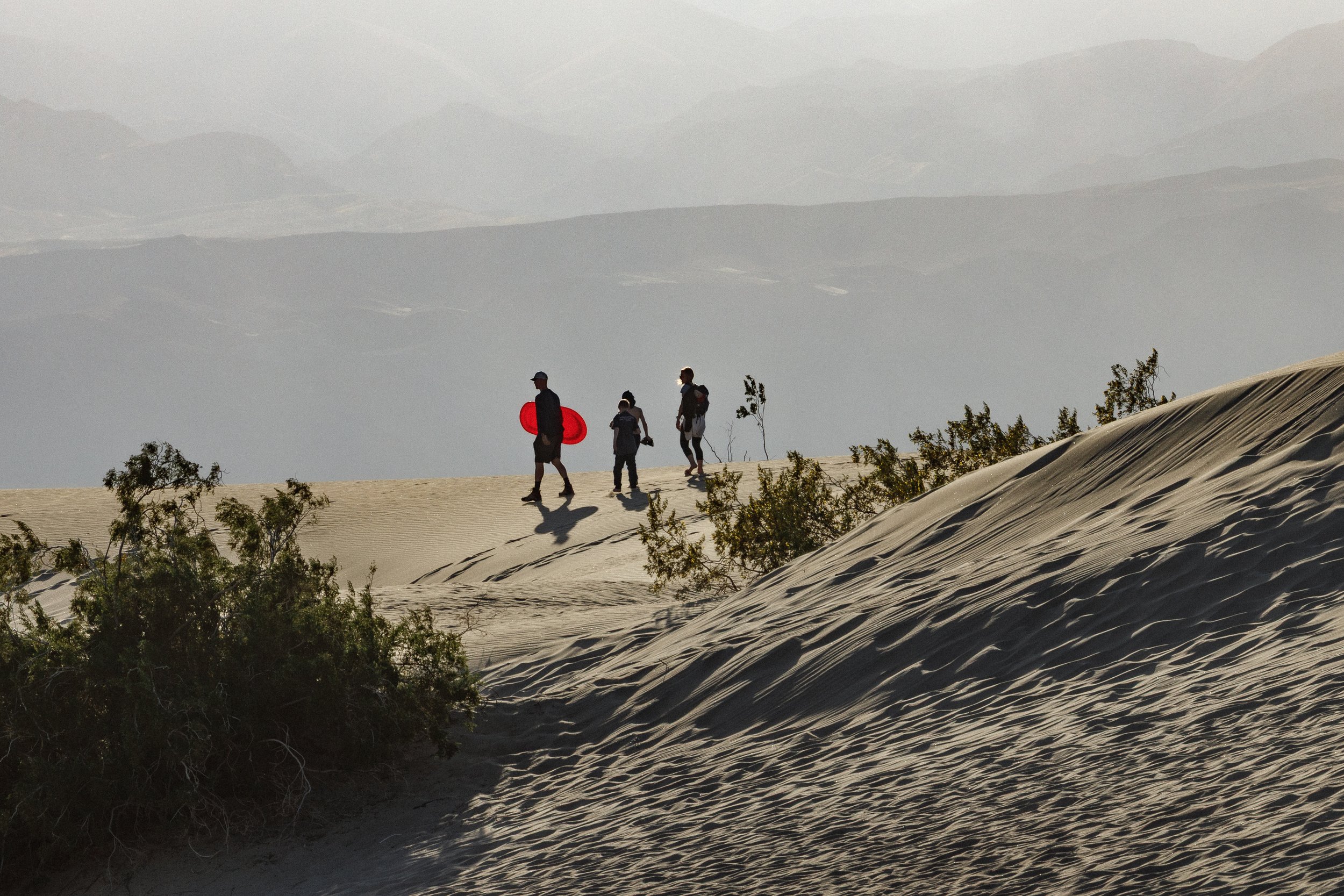 A group of four people walking on sand dunes in a desert landscape with some sparse vegetation and mountains in the background.