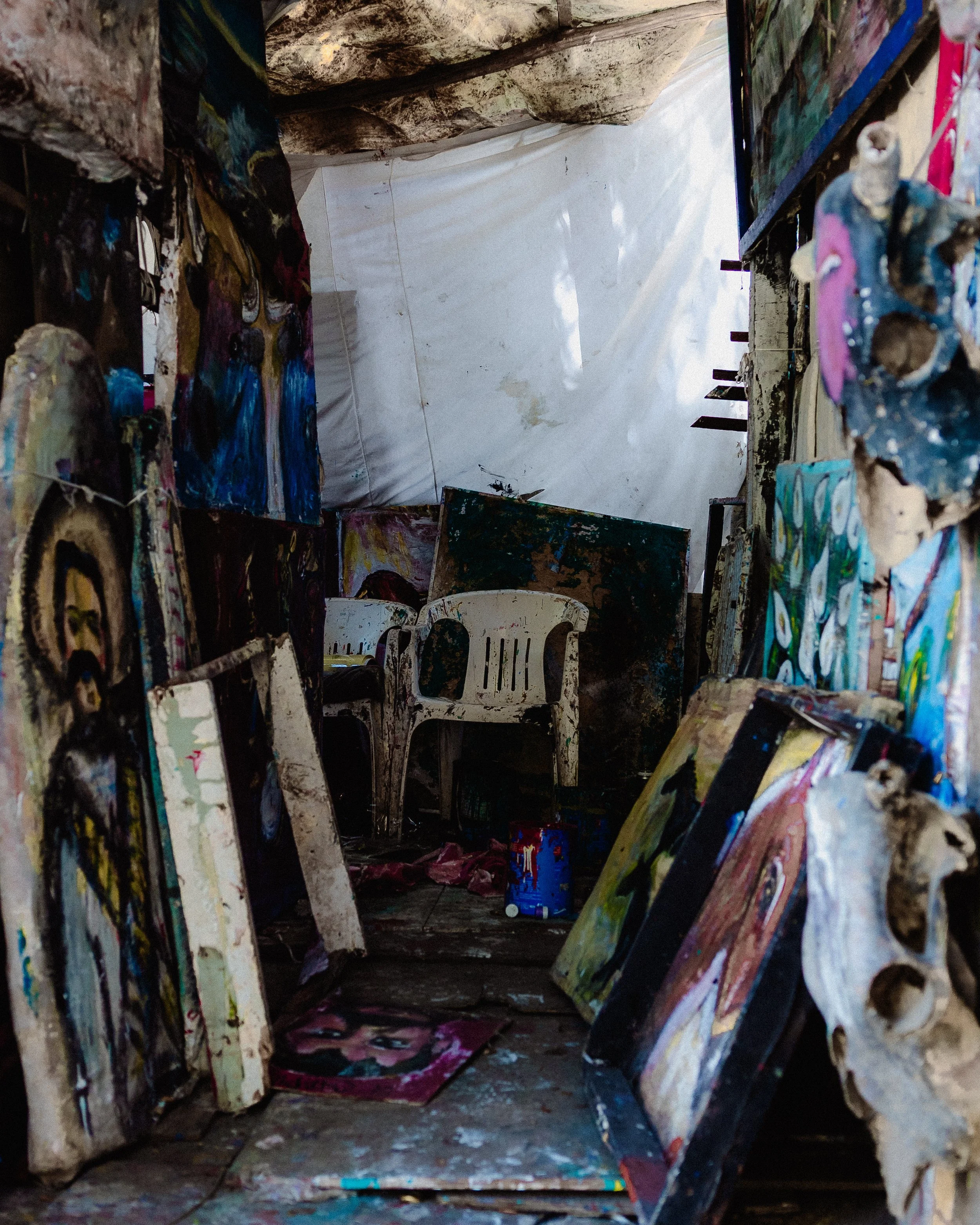 An art studio with paintings stored on the walls and floor, a white plastic chair, and supplies like paint cans, with natural light coming from outside.