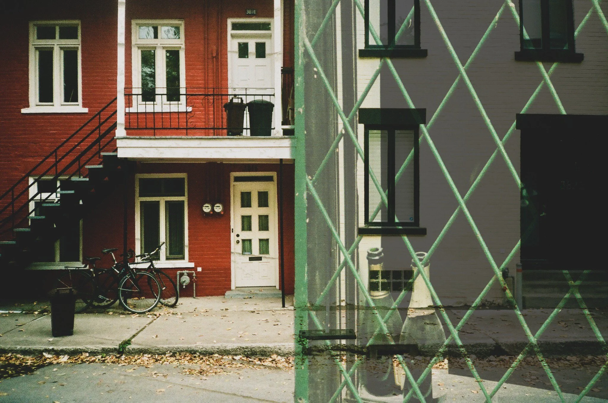 Shot of a residential building viewed through a chain-link fence. The building has a red exterior with white window frames and a white door, with bicycles parked outside and trash bins on the balcony.