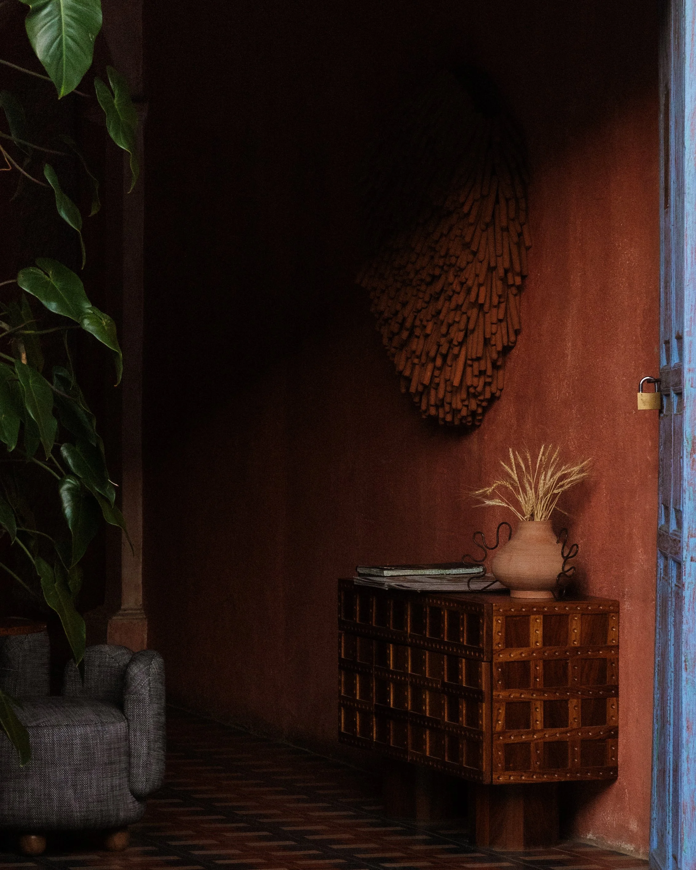 Interior of a cozy room with a plaid sofa, a wooden chest with multiple small drawers, a large ceramic vase with dried grasses, a leafy green plant on the left, and a wall mounted with a moss wall art piece of a heart shape.