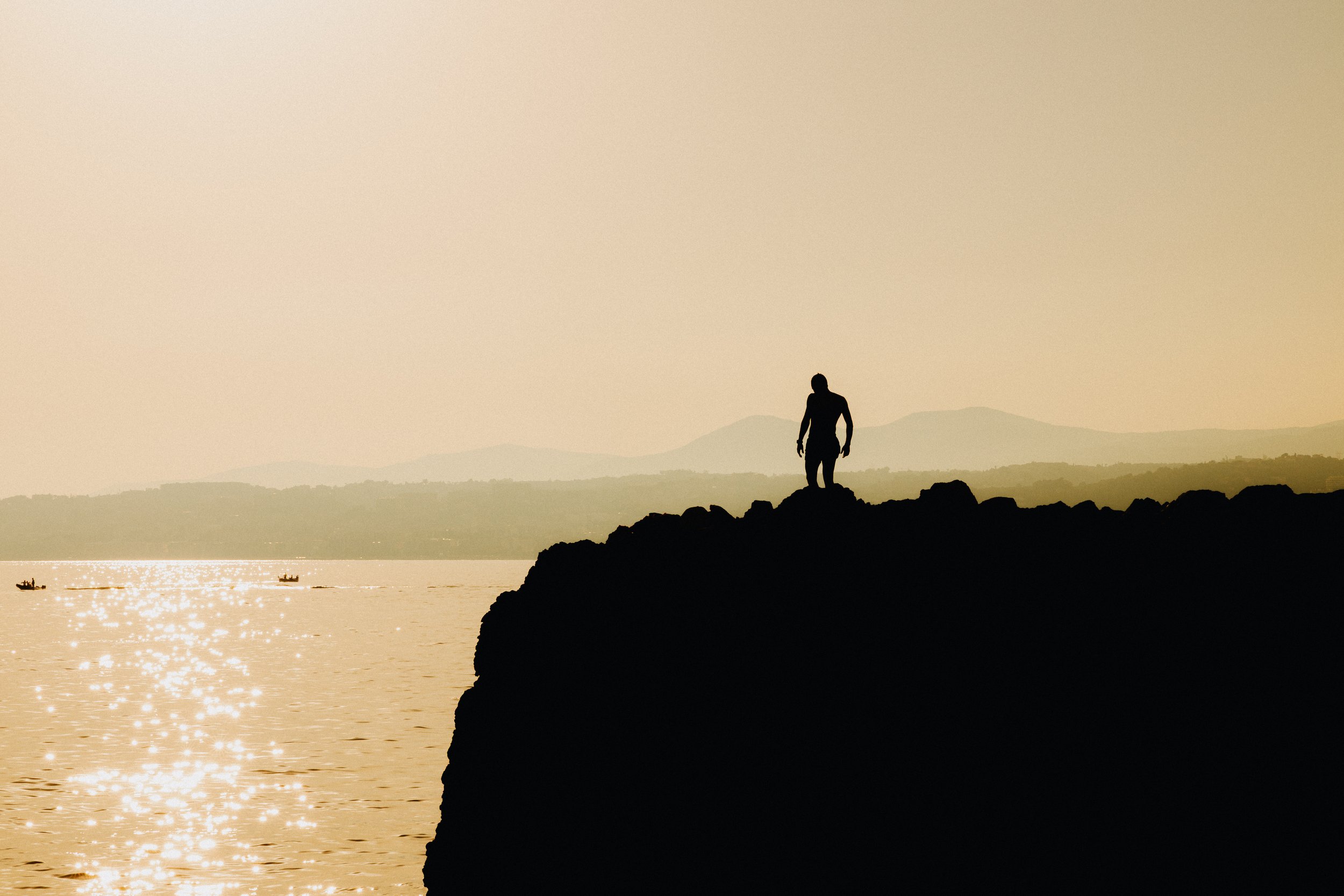 A silhouette of a person standing on a rocky ledge overlooking a body of water during sunset or sunrise, with boats visible on the water and mountains in the distance.