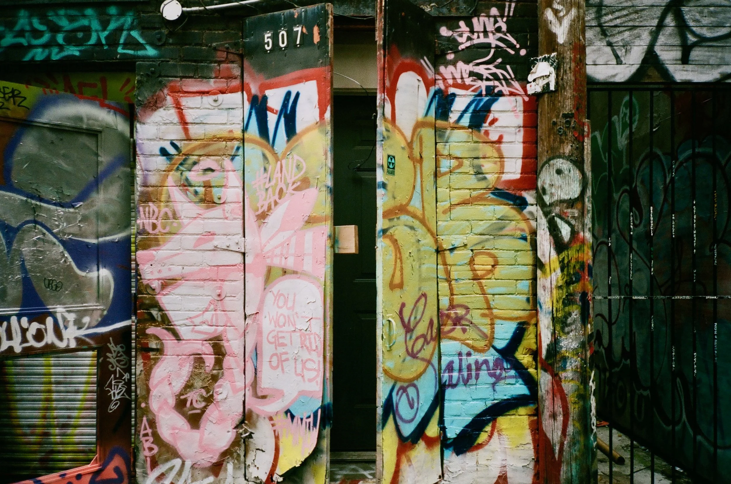 Close-up of a graffiti-covered wooden and metal surface, including a door, with vibrant spray paint tags and designs in various colors.
