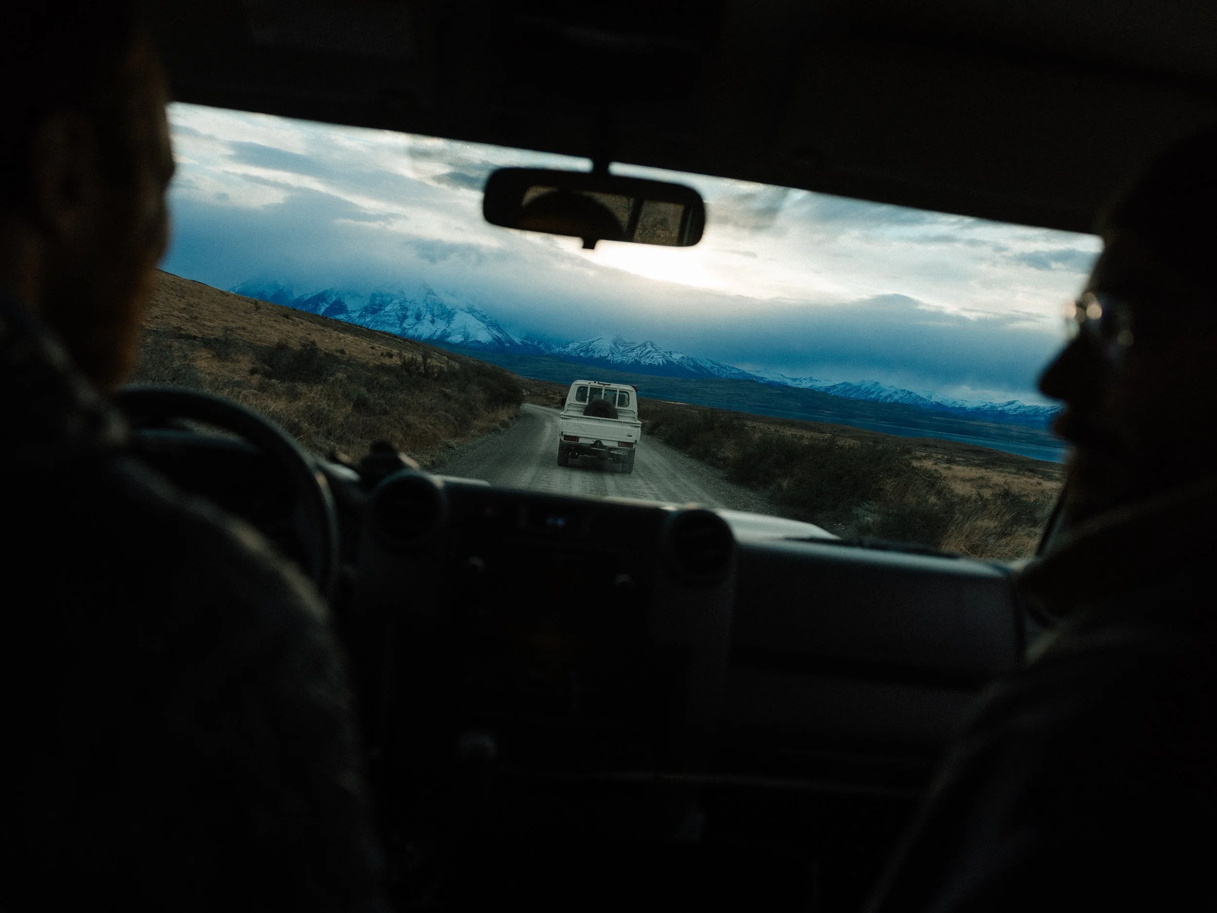 View from inside a vehicle showing a dirt road and a white vehicle ahead, with snow-capped mountains and cloudy sky in the background.