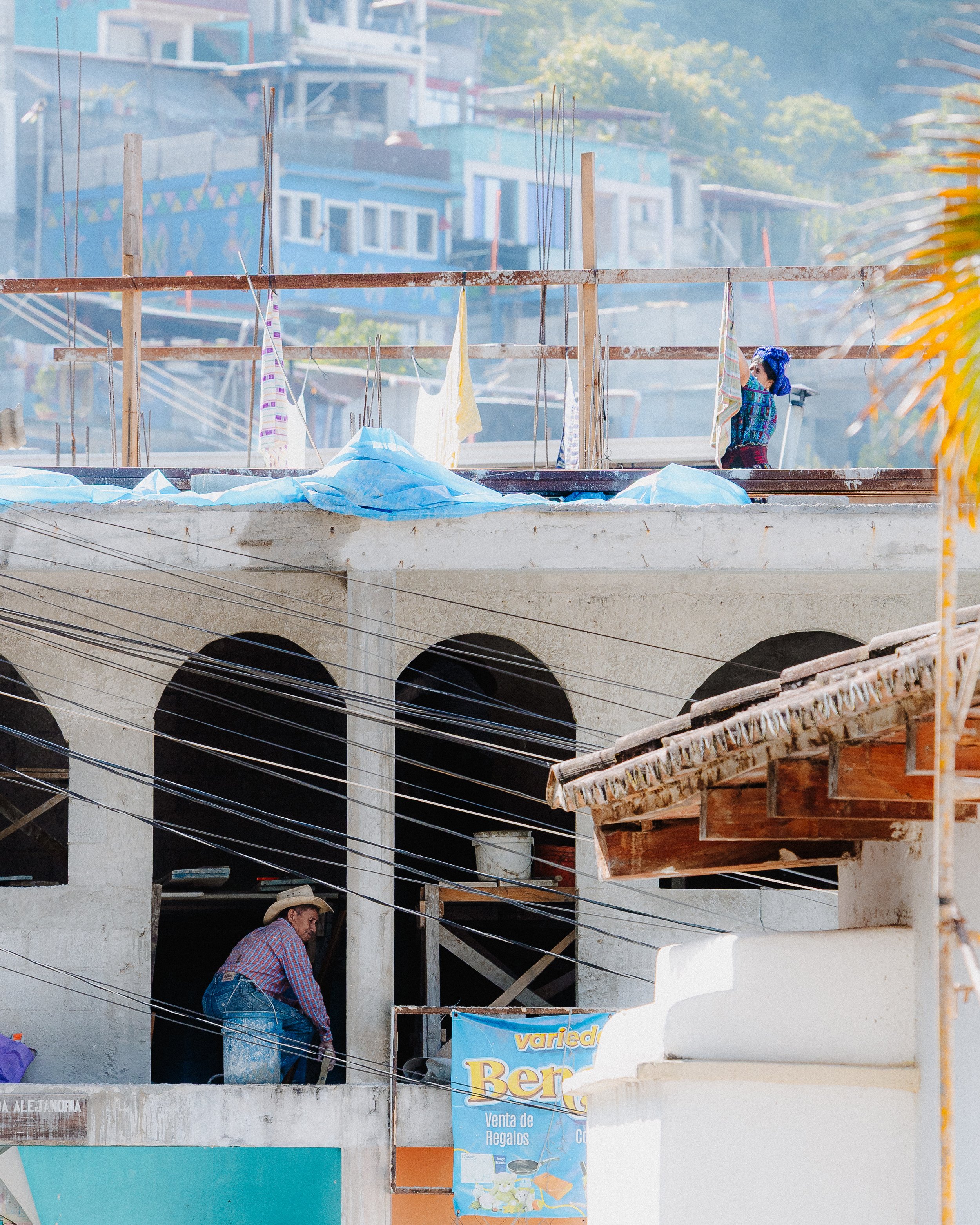 A man in a plaid shirt and jeans working on a building under construction, with wooden beams and laundry hanging on cords. Multiple apartment buildings in the background and a blue advertising banner in the foreground.