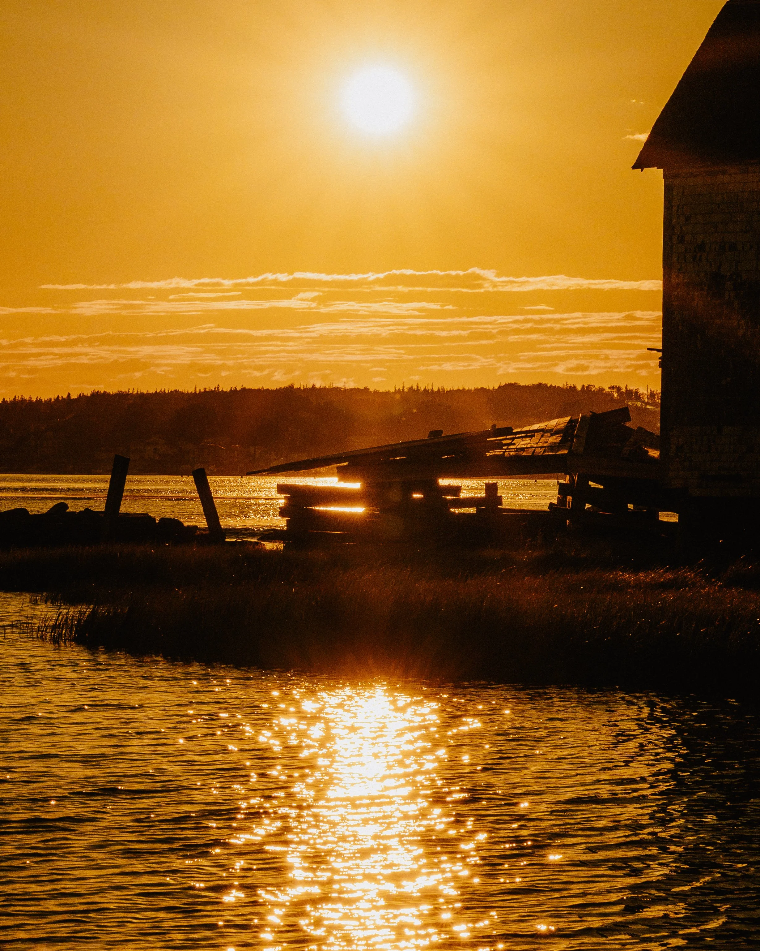 Sunset over a body of water with reflections, a silhouette of a house on the right, and a wooden dock in the foreground.