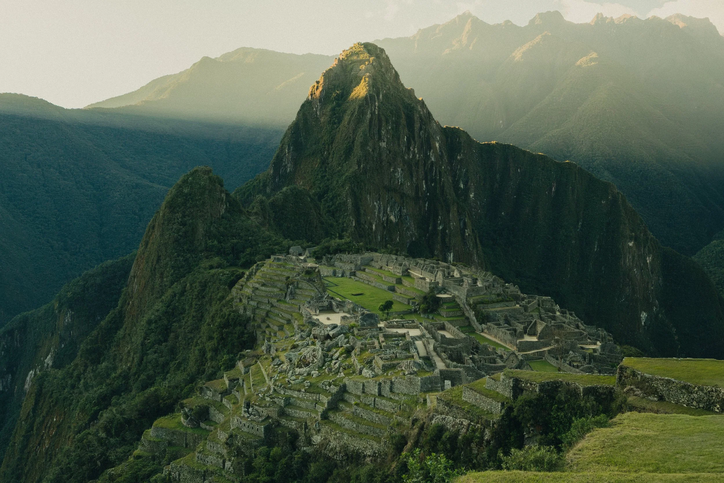 Ancient Incan ruins of Machu Picchu with stone terraces and buildings, surrounded by lush green mountains and a towering peak in the background.