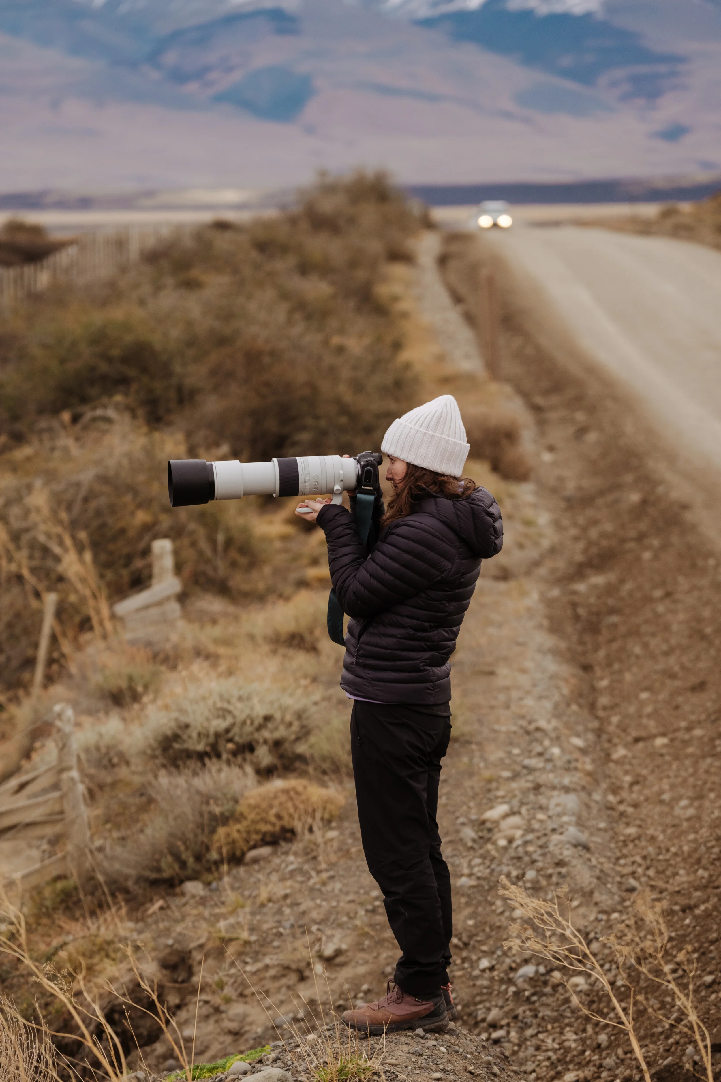 Woman in a white knit hat and black jacket standing outdoors on rocky, dry terrain during daytime, holding a large white telescope with black accents, with mountains and a winding road in the background.