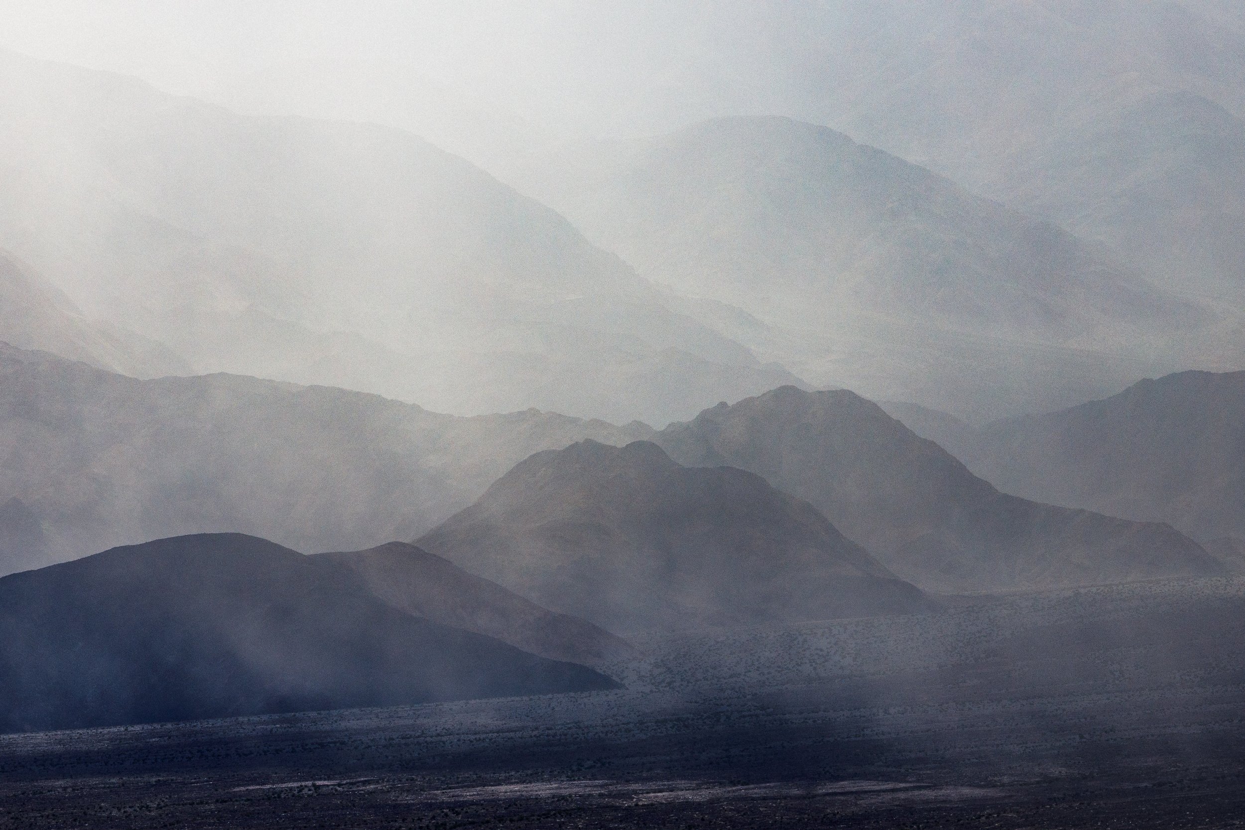 A desert landscape with multiple layers of mountains in shades of blue and gray, under a cloudy sky.