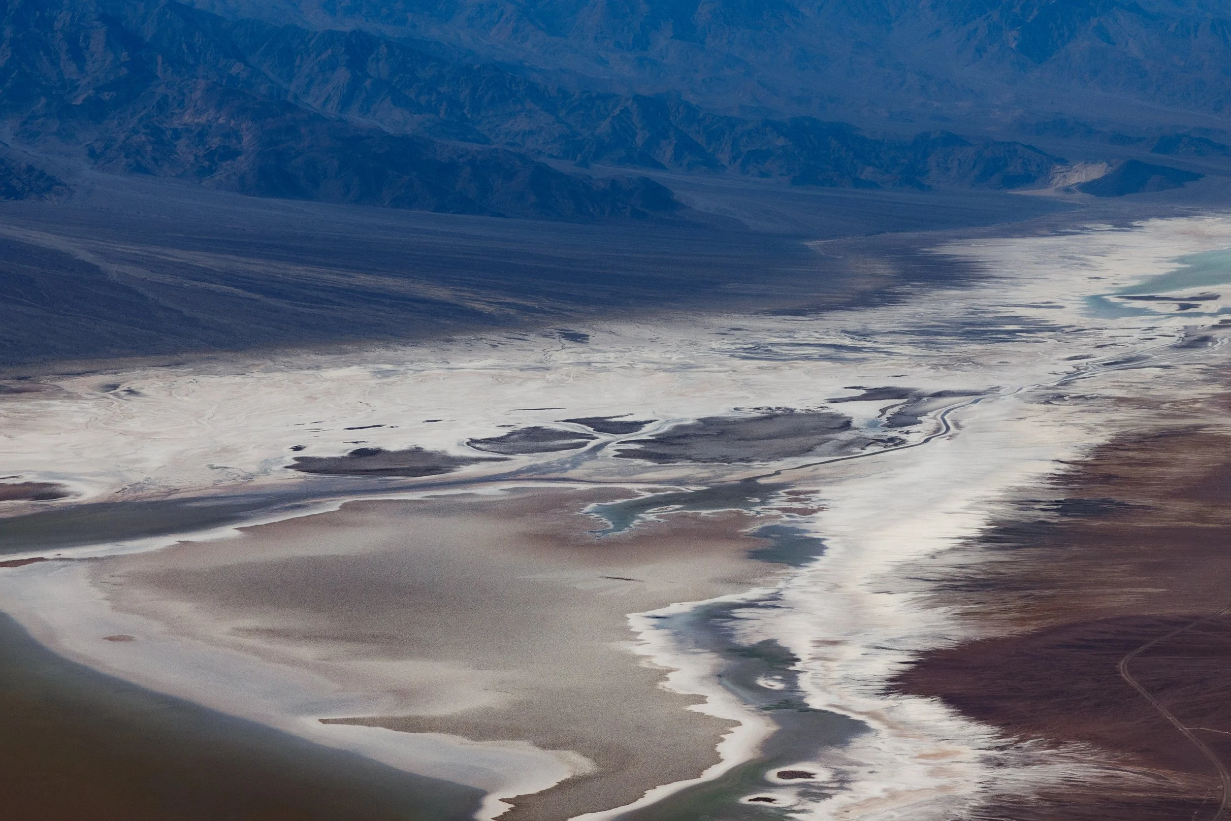 Aerial view of a flat, dry landscape with salt flats, mountains in the background, and a winding road visible in the lower right corner.