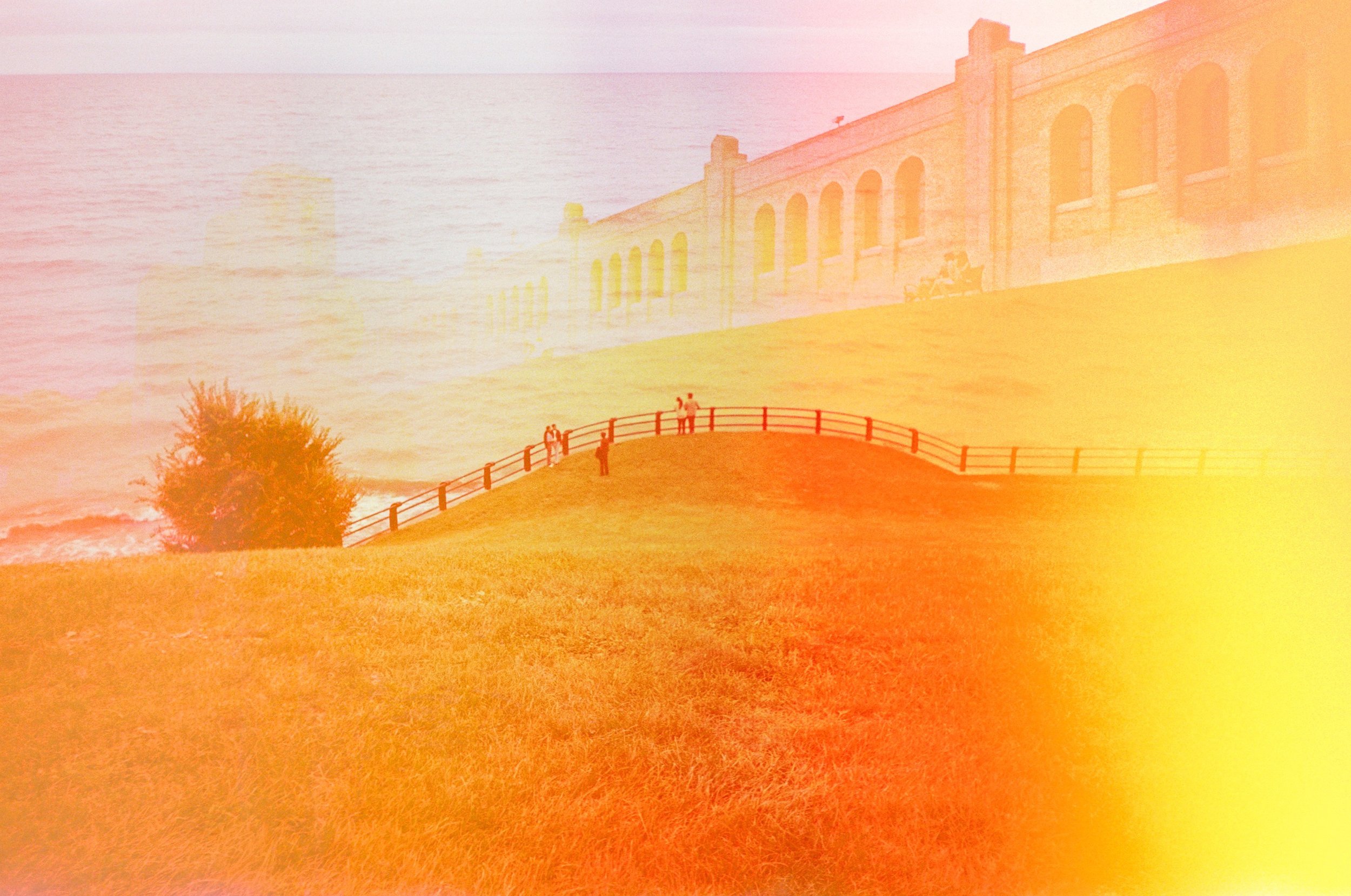 A park with a grassy hill, a wooden fence, a tree, and a large brick building in the background, with a sunset over the water.