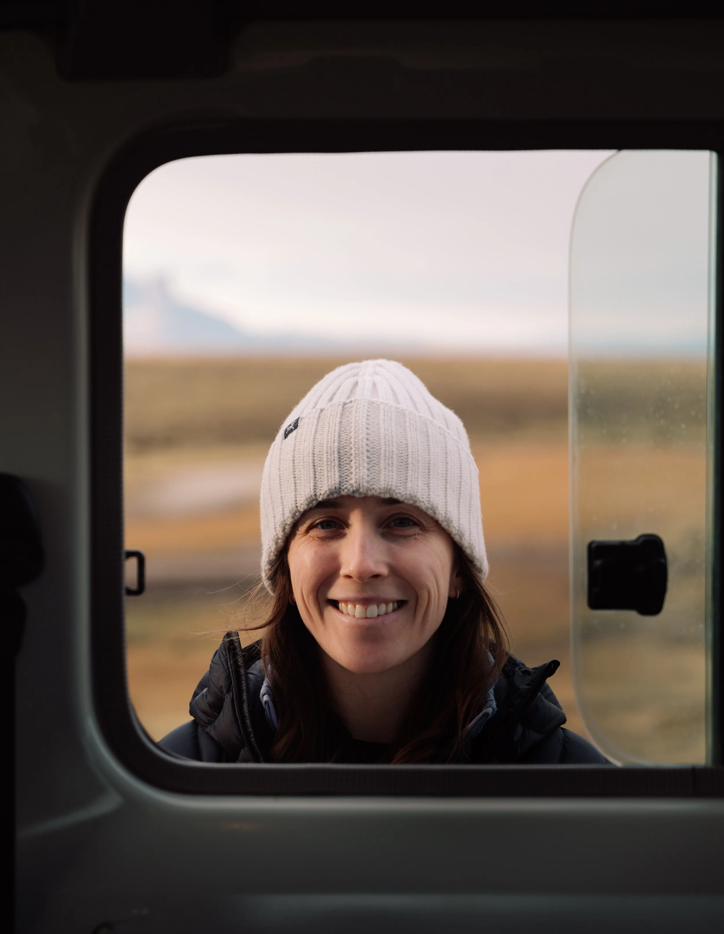 A woman smiles while looking through a window at a landscape of open fields and cloudy sky. She is wearing a cream knit beanie and a dark jacket.