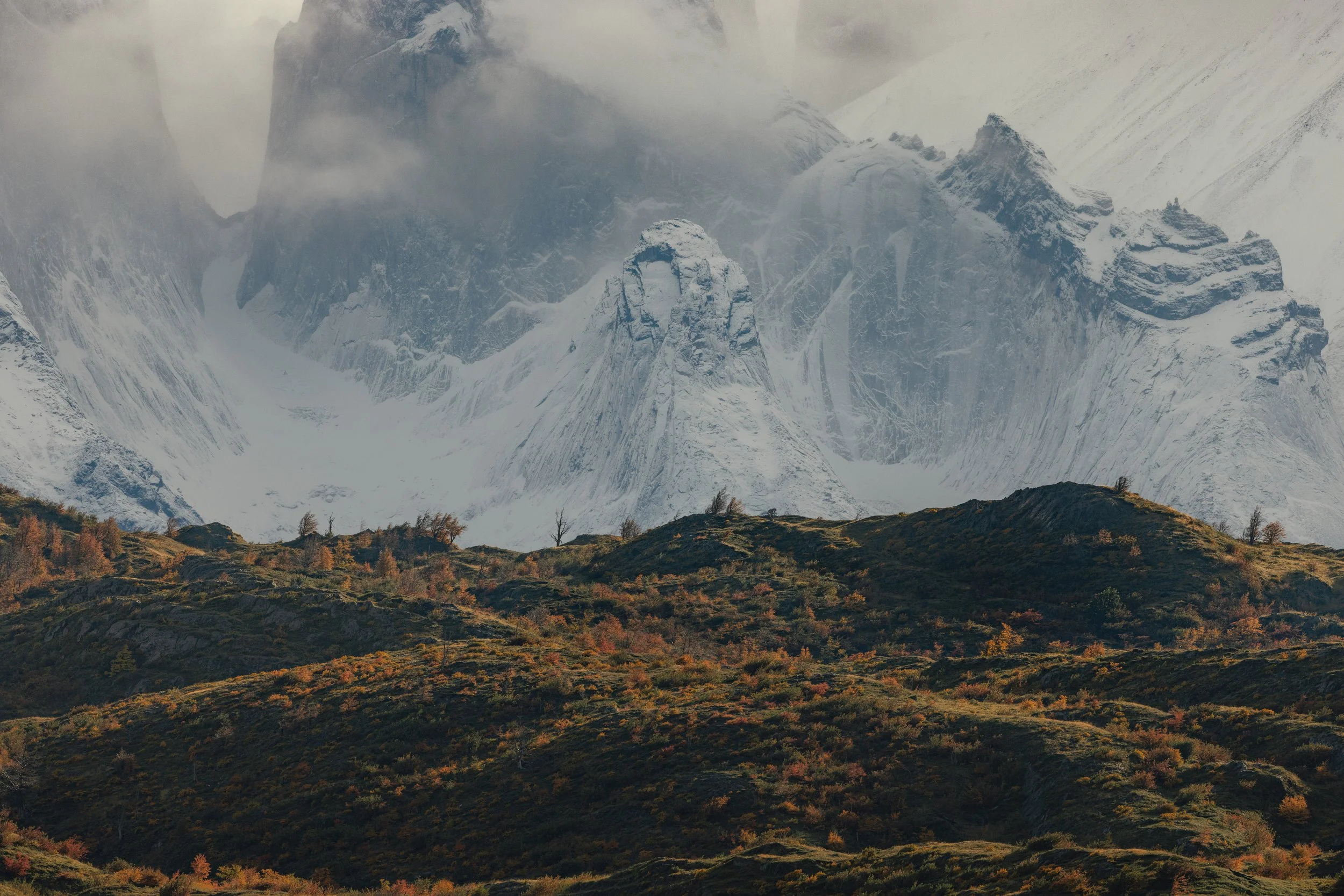 Snow-covered mountain range with steep rocky peaks and a valley at the base with sparse trees and vegetation, under cloudy sky.