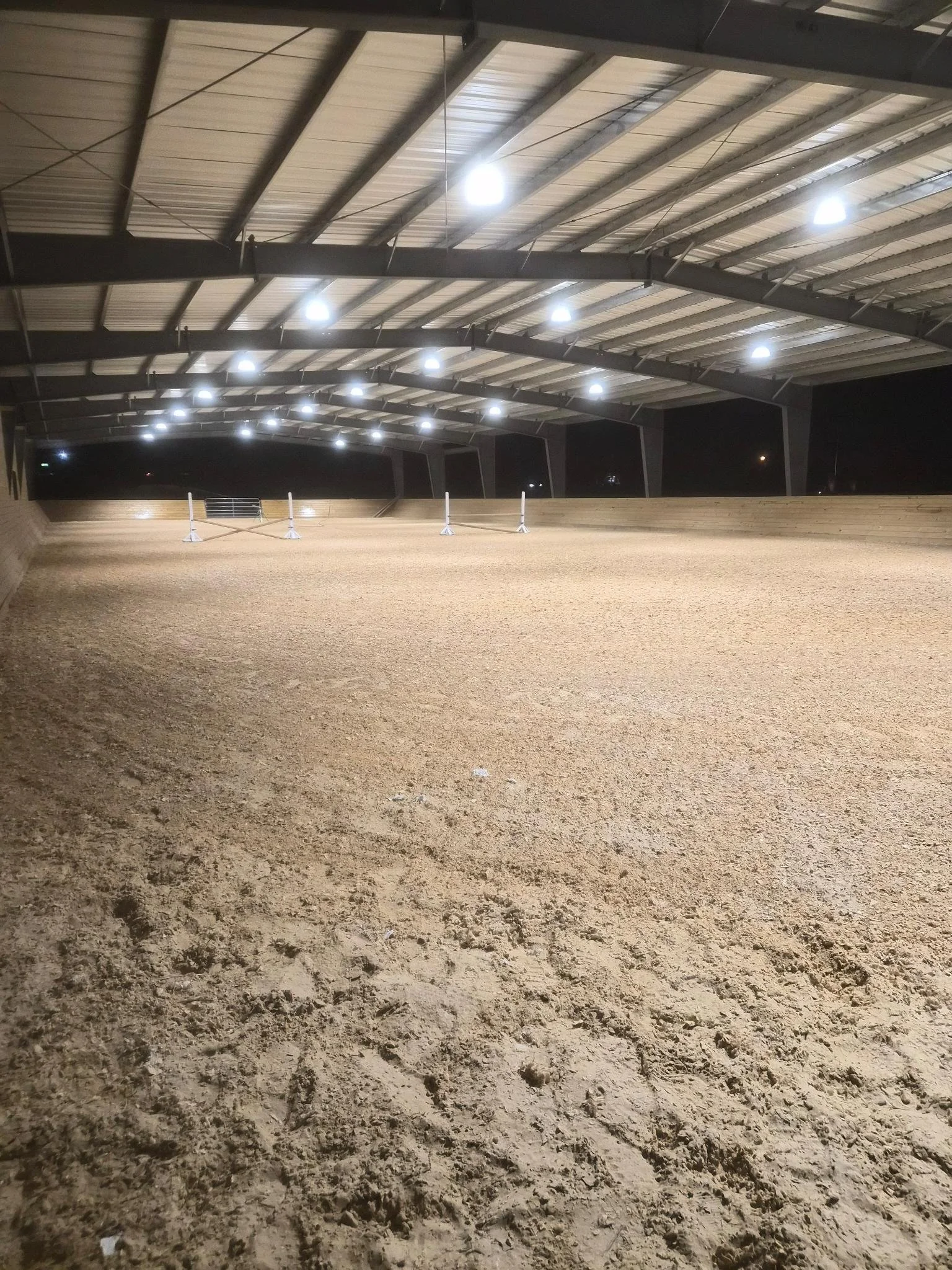Our riding arena with competition footing, wooden fences, and overhead bright lighting in a covered structure.