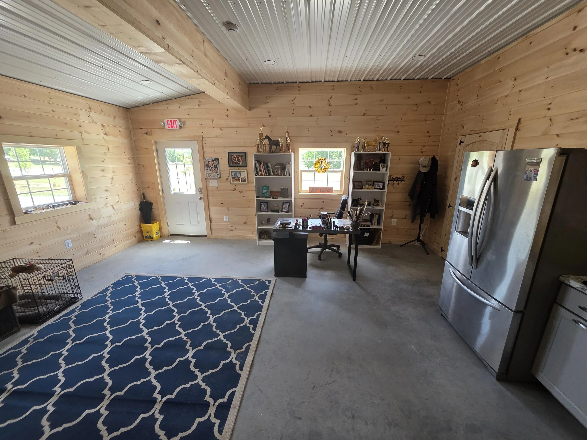 Boarding lounge area with wooden walls and ceiling, two windows, a door, a fridge, a bookshelf, a desk with office supplies, and a blue and white patterned rug.