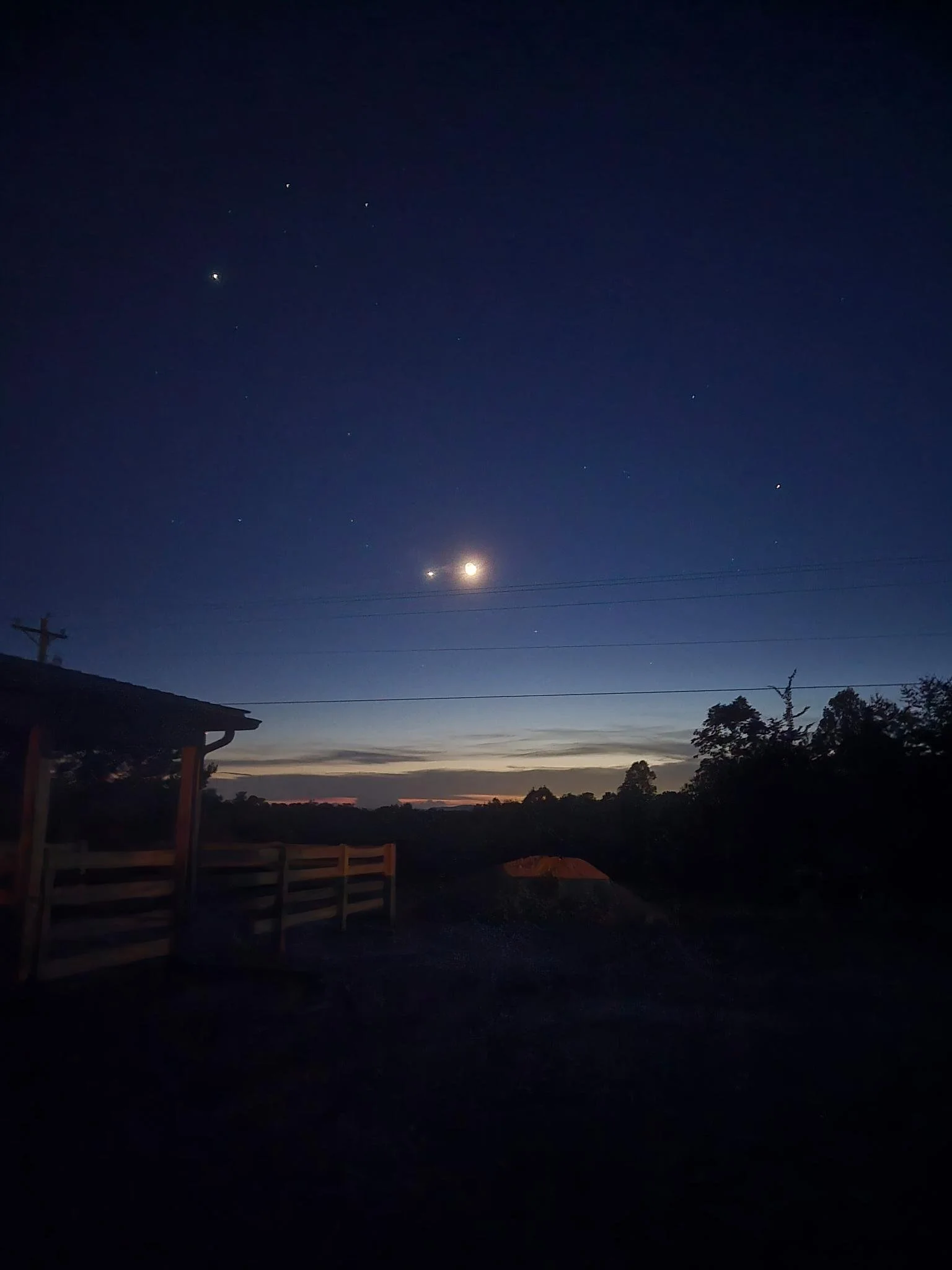 Night sky over the farm with visible planets, stars, and the moon above a rural landscape with trees and a wooden structure.