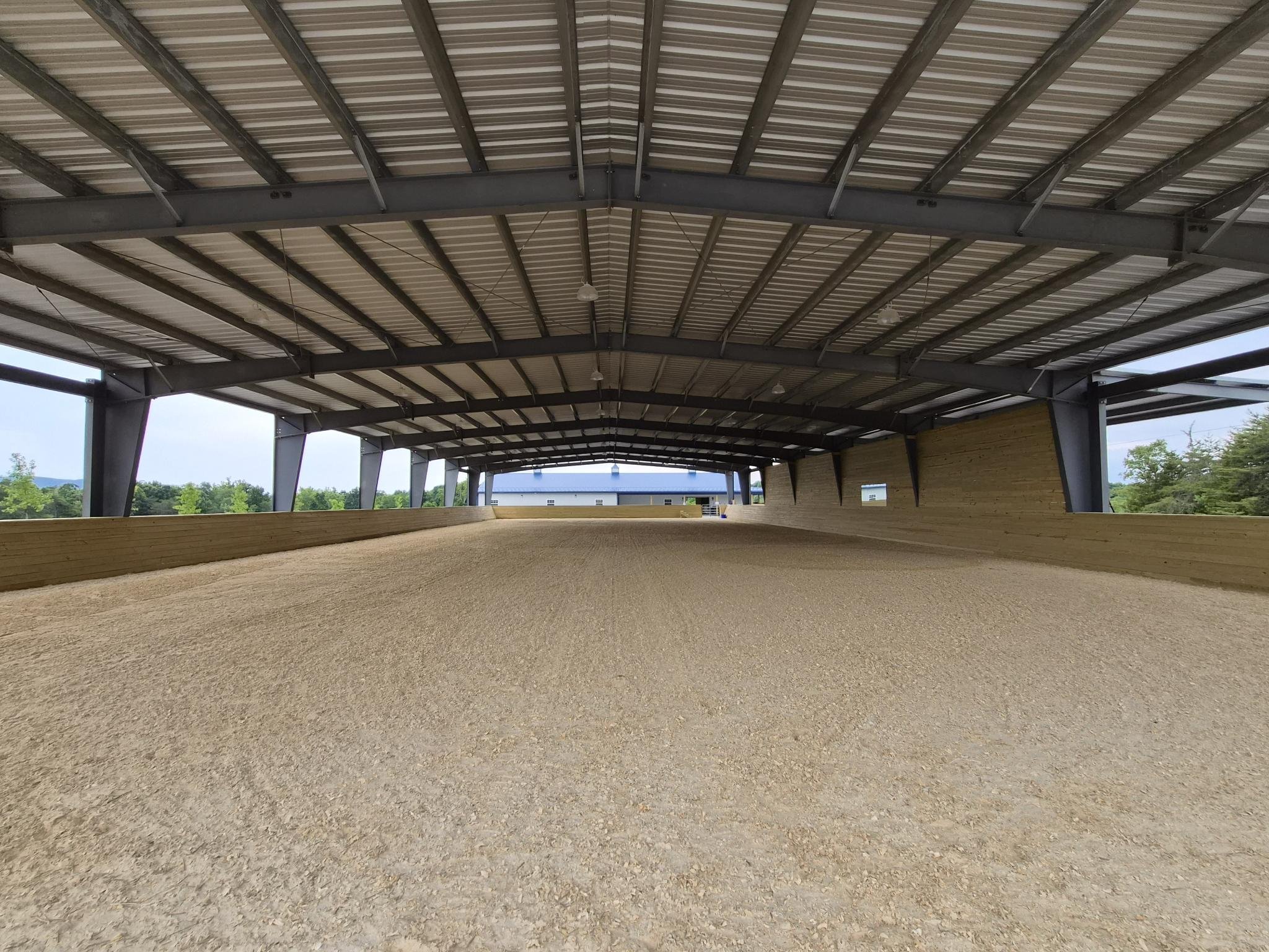 Indoor equestrian riding arena with a dirt floor, surrounded by wooden barriers and supported by a metal roof structure.