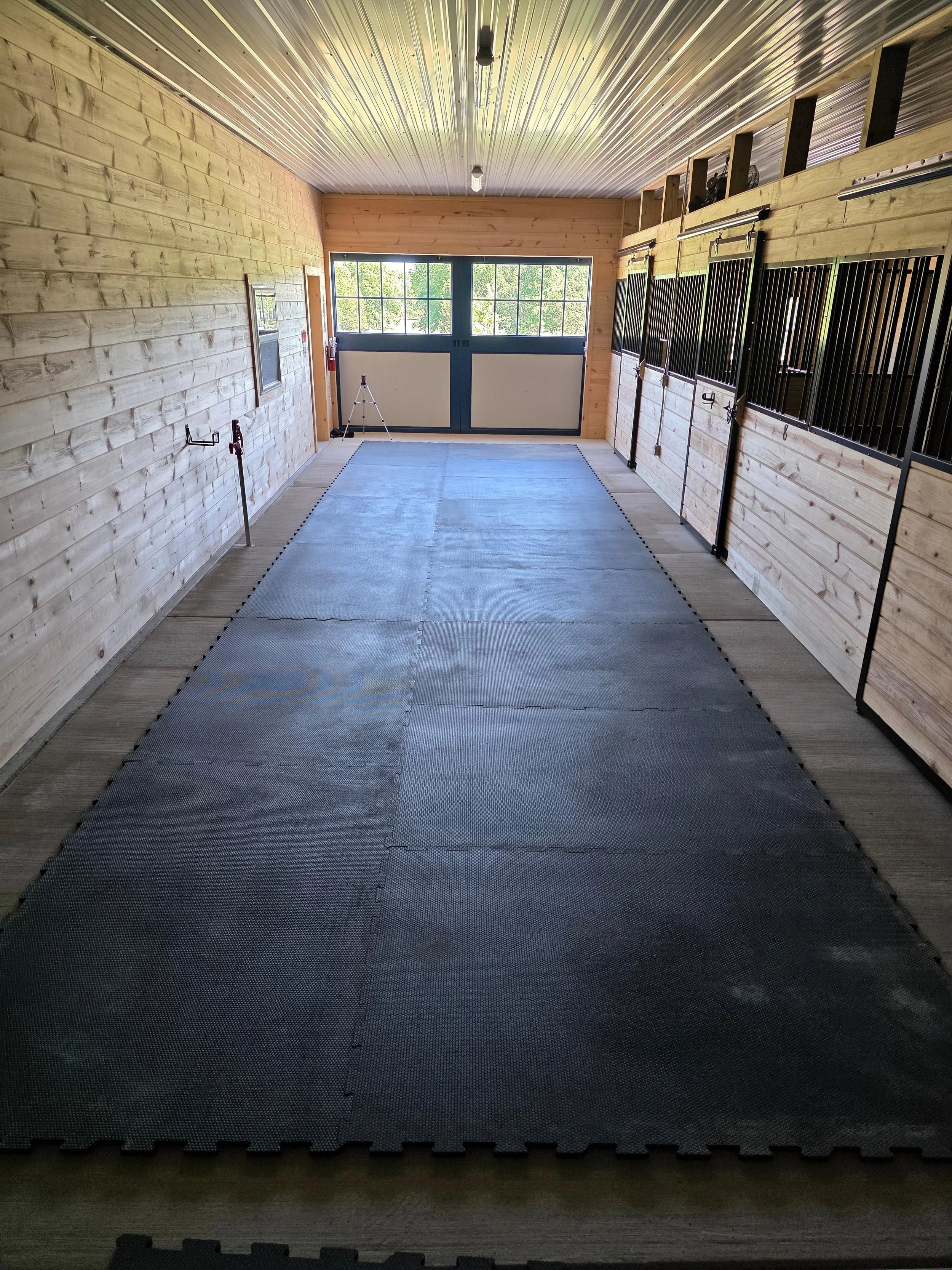 The main aisle in the stable with black metal bars on the stalls, a large window at the end, and a rubber mat on the floor.