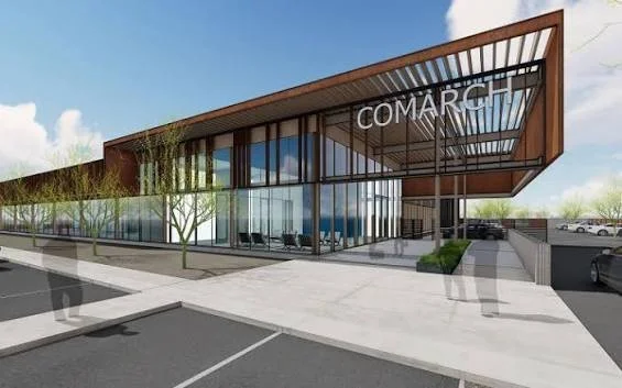 Modern commercial building with glass facade and wooden accents, labeled COMARCH, located in a parking lot with a few cars and trees.