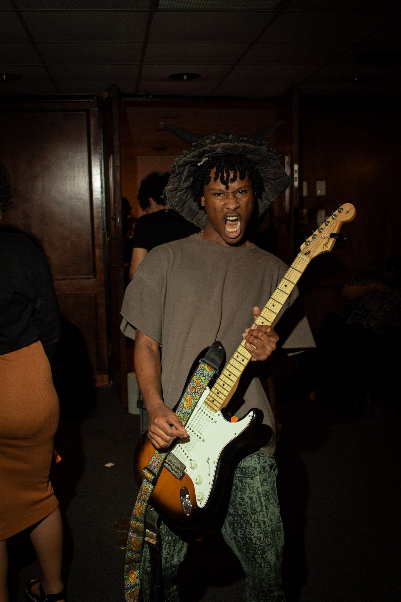 A person with dark skin and curly hair, wearing a large hat and patterned pants, passionately singing and playing an electric guitar with a colorful strap in a dimly lit indoor setting.