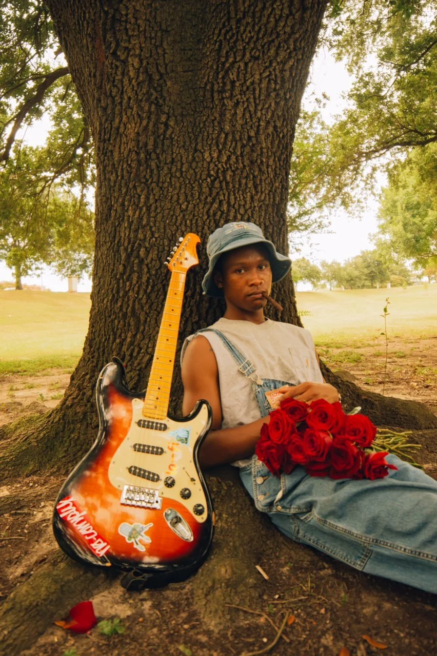 A young boy sitting under a large tree, holding a bunch of red roses and a dollar bill, with a guitar leaning against the tree beside him. The boy is wearing a bucket hat and overalls, and has a cigar in his mouth.