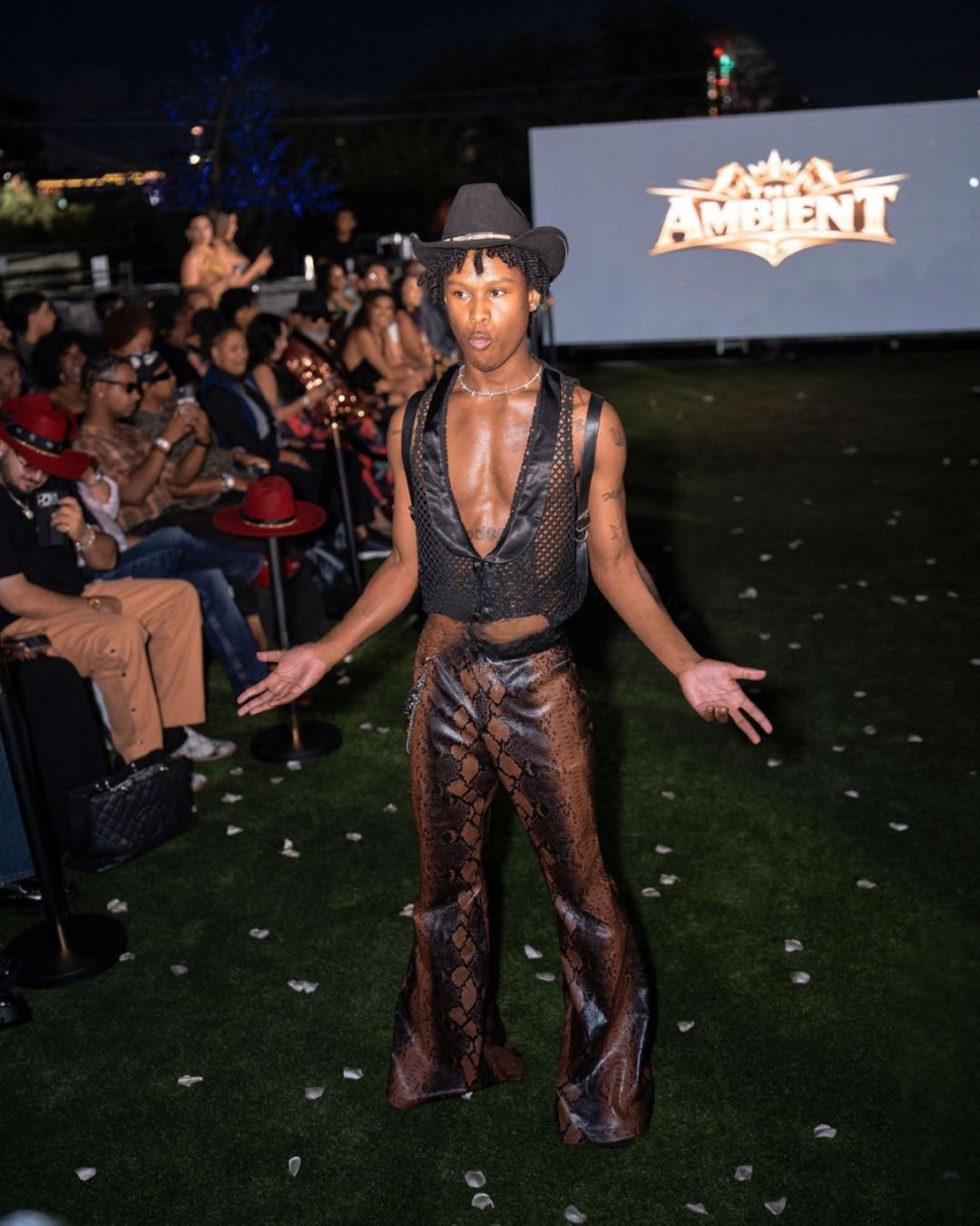 A person walking on a runway at an outdoor fashion event during the night. The person is wearing a black wide-brimmed hat, a black netted sleeveless top with a deep neckline, and shiny brown snakeskin-patterned wide-leg pants. There is a large screen in the background displaying the word "AMBIENT." Audience members are seated on either side of the runway.