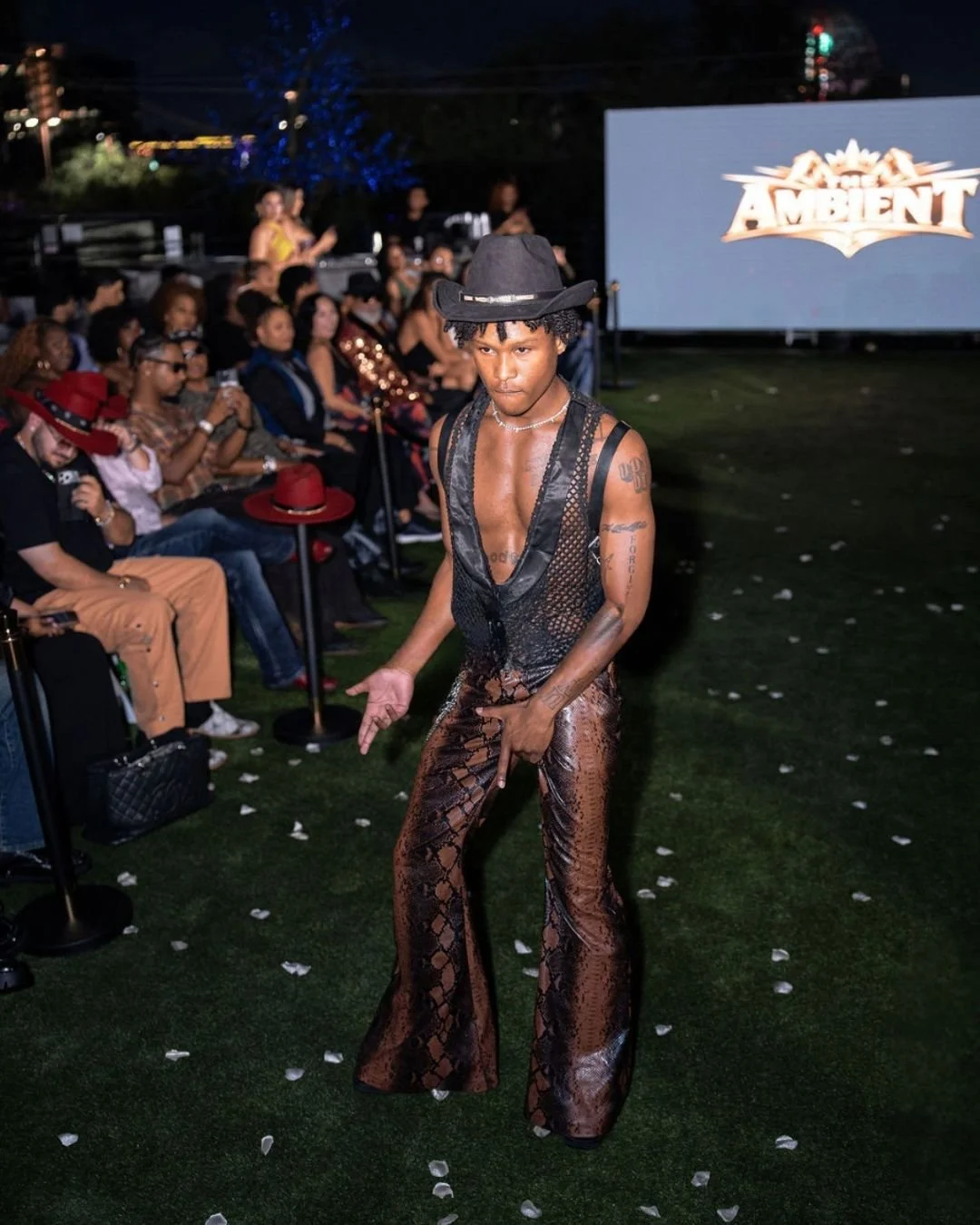A young man dancing at a fashion show wearing a cowboy hat, lace-up brown snakeskin pants, and a sleeveless black top, with an audience seated behind him watching.