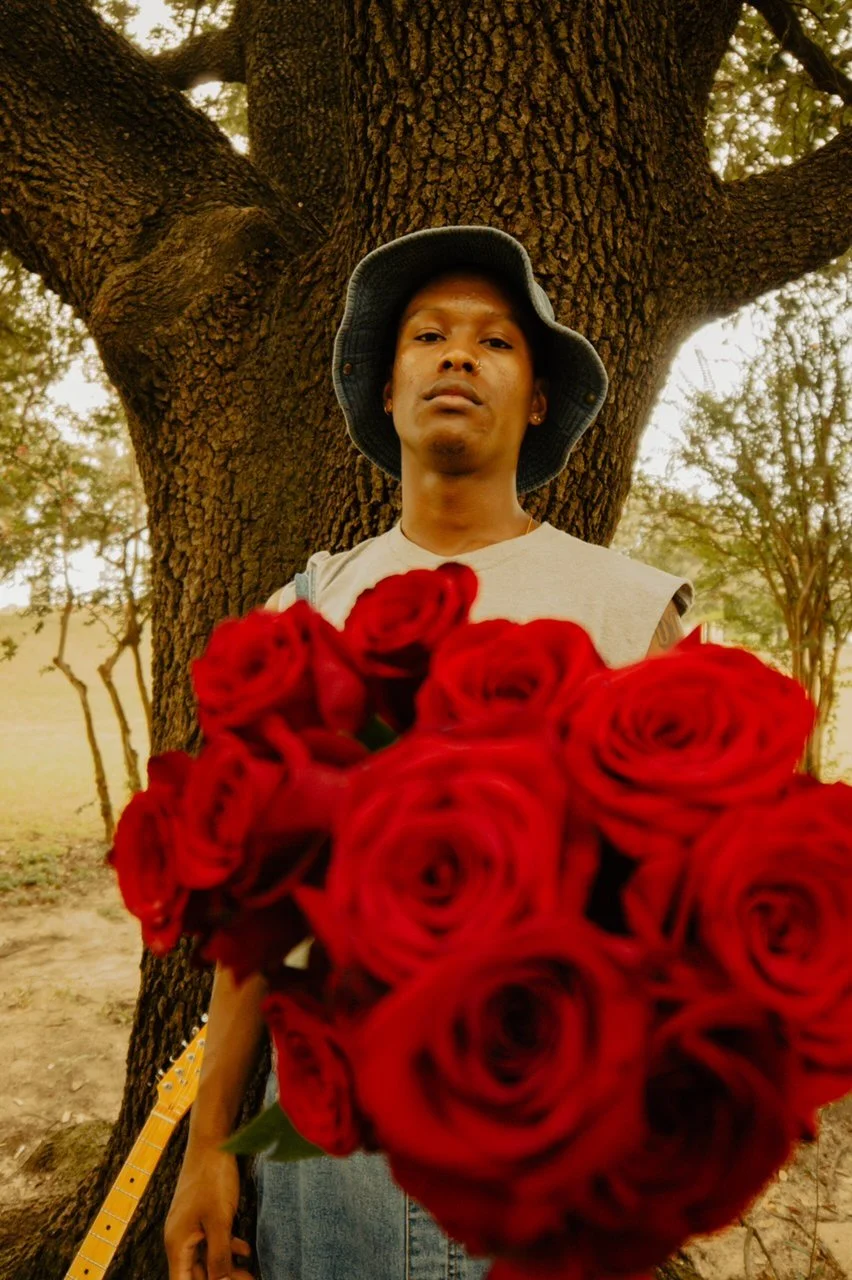 A person standing outdoors in front of a large tree, holding a big bouquet of red roses, wearing a bucket hat and a sleeveless shirt.