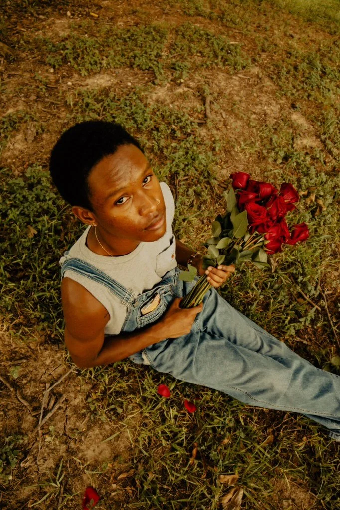 Young woman sitting on ground holding a bouquet of red roses in an outdoor grassy area.