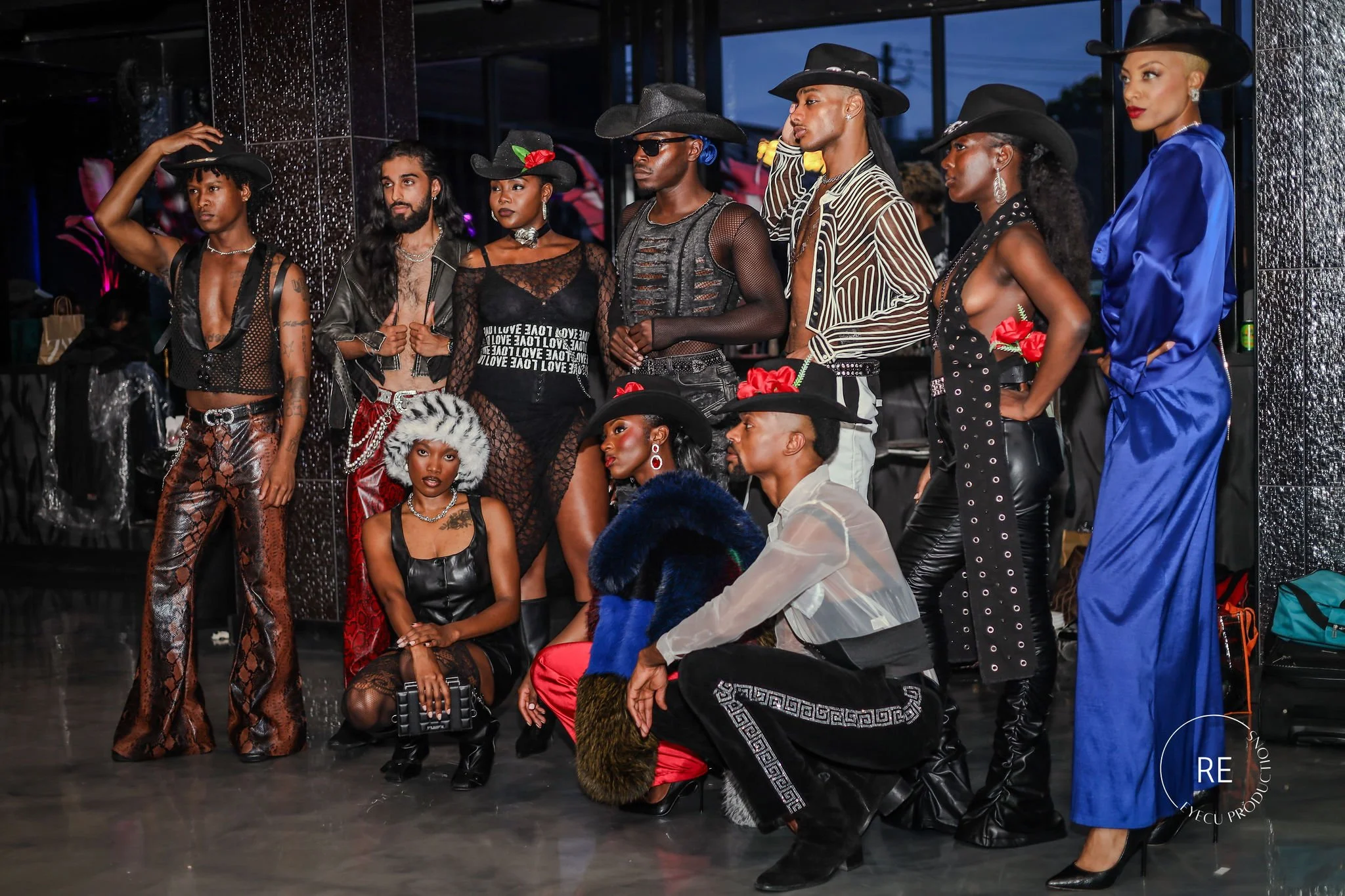 A group of eight fashionably dressed models, six women and two men, posing backstage at a fashion show. They wear bold, stylish outfits with cowboy and western influences, including hats, leather, lace, and metallic details. Some women have accessories like earrings and flowers in their hair. They are standing or sitting in front of a mirror and a black reflective surface, with a nighttime city view visible through the windows.