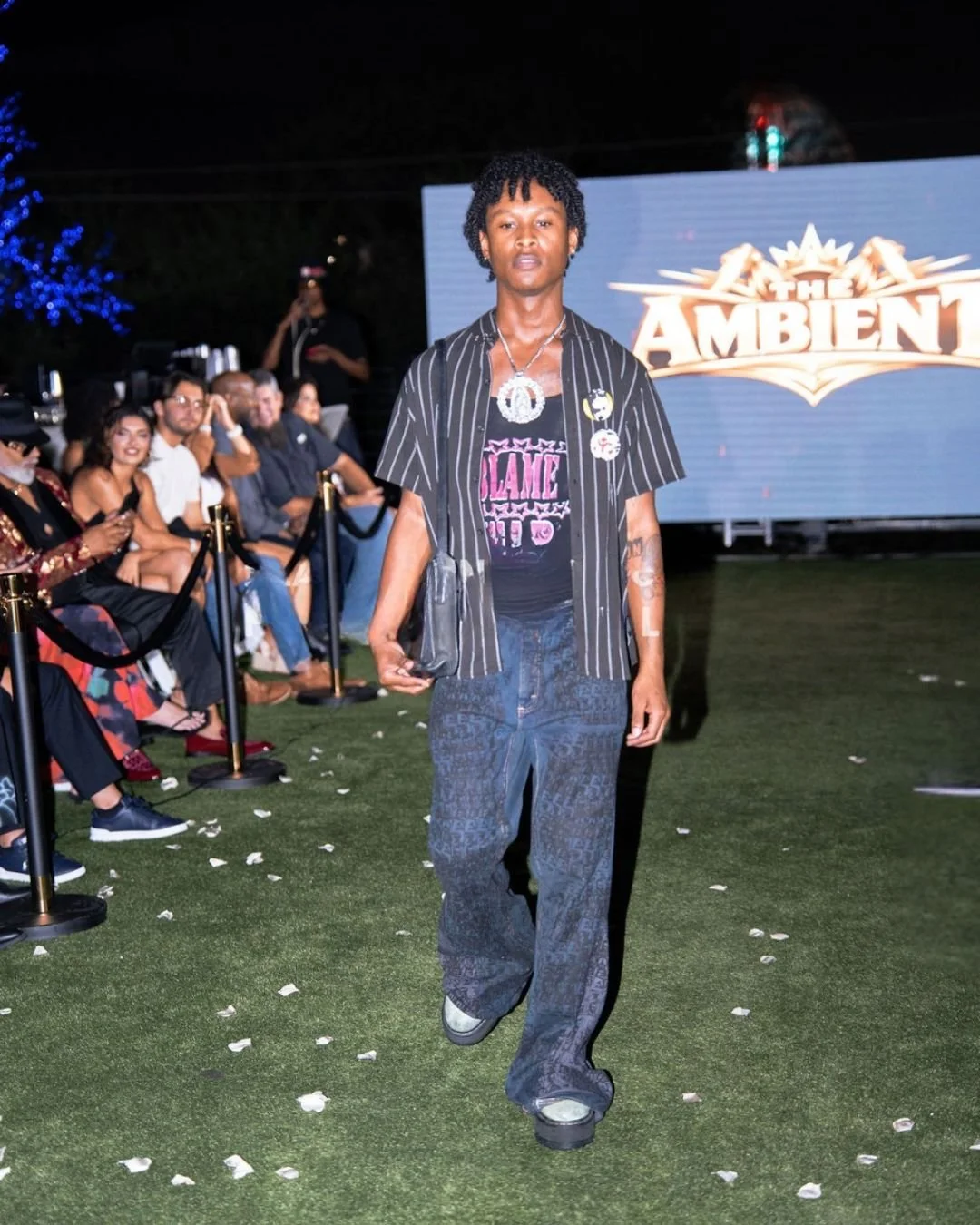 A person with curly hair walking on a green carpet at an outdoor event at night, with an audience seated behind a black barrier. There is a large screen with the words 'The Ambient' in the background.