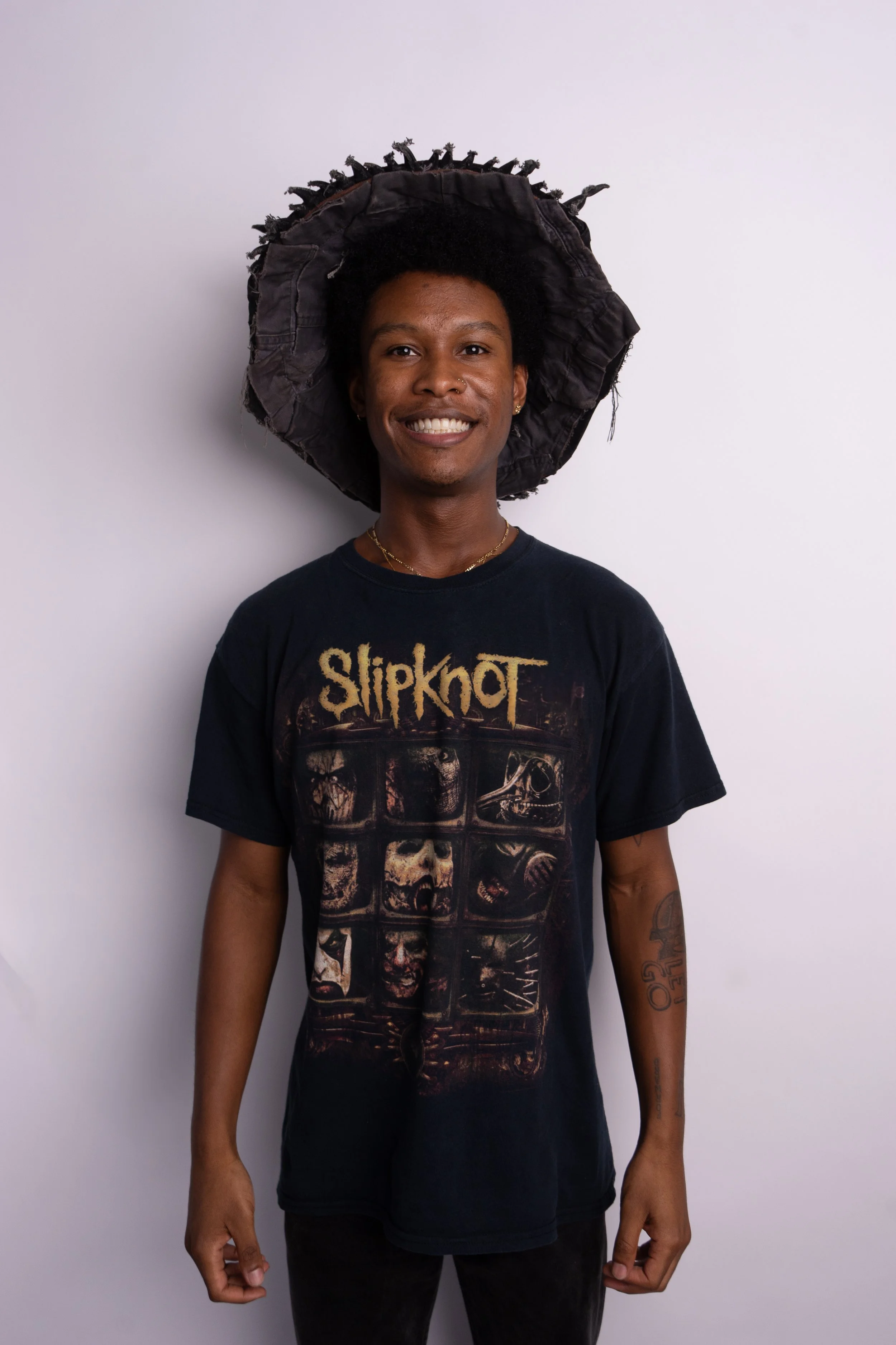 A smiling person with dark skin and short curly hair, wearing a large black wide-brimmed hat made of fabric with frayed edges, a black Slipknot band T-shirt with a graphic design, and black pants, standing against a plain light gray background.