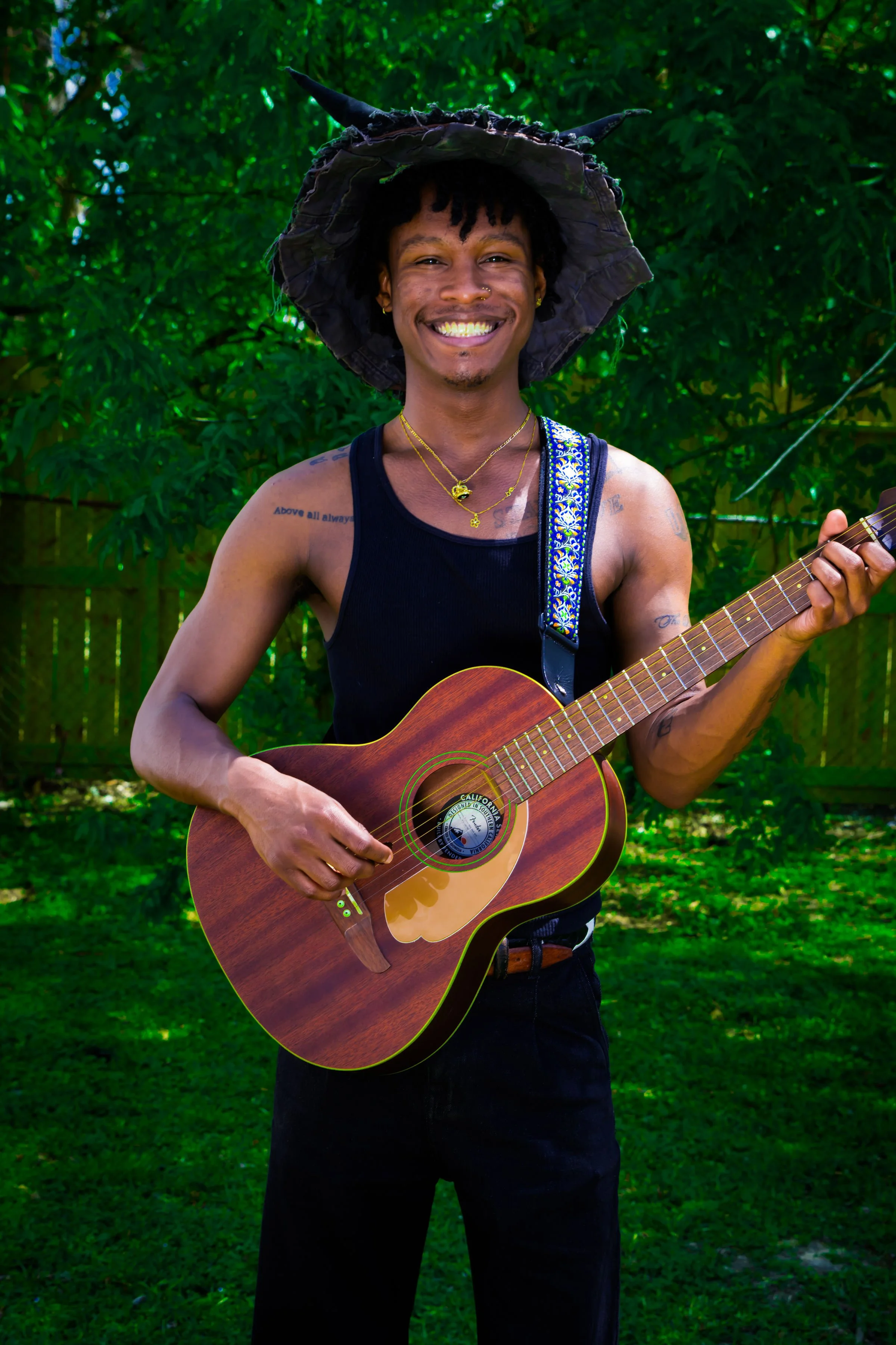 Smiling man with curly hair wearing a black tank top and black pants, holding an acoustic guitar, standing outdoors with green foliage in the background.