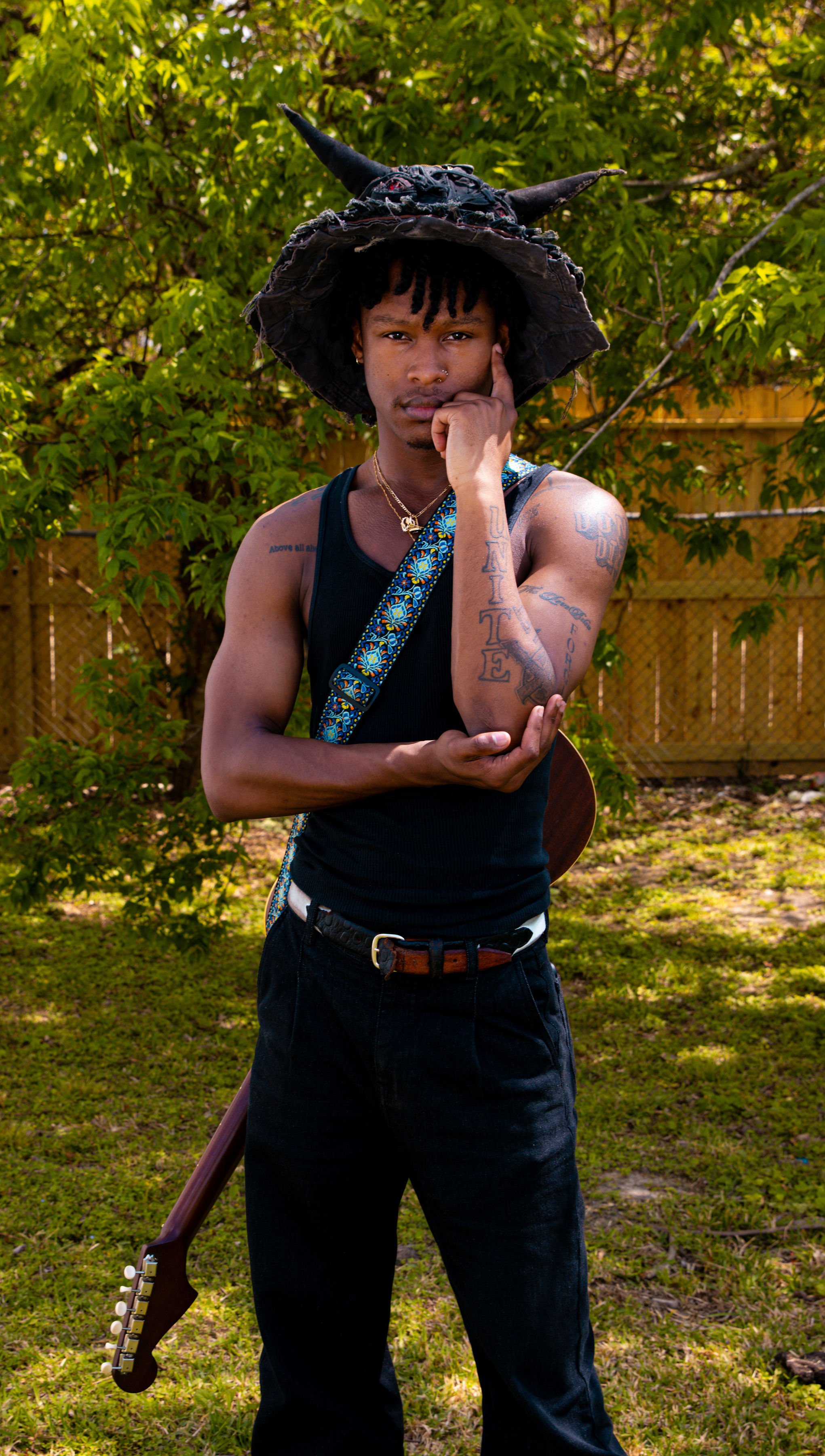 Young man with tattoos on his arms, wearing a black tank top and a large, black, tattered hat with horns, holding a guitar and standing outdoors in front of trees and a wooden fence.