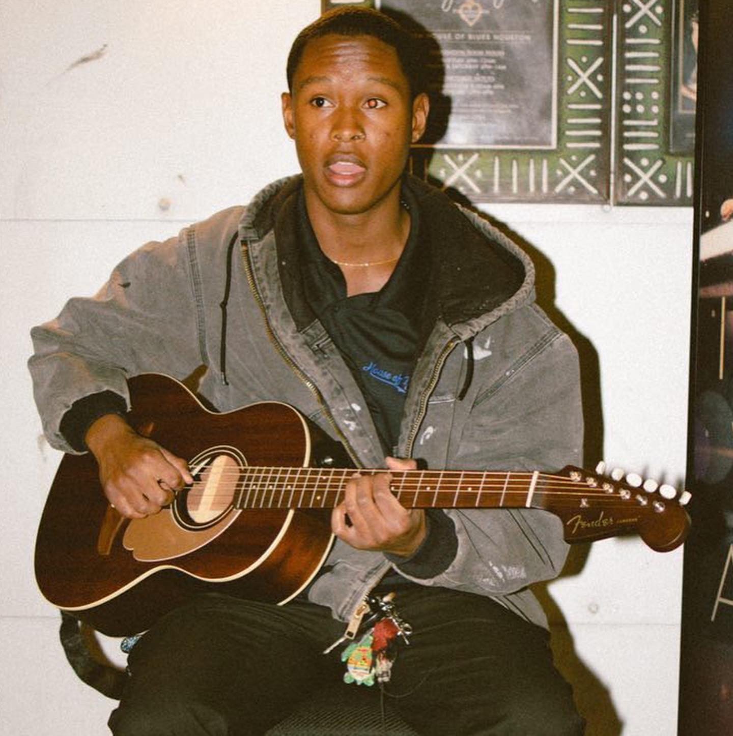 A person with dark hair playing an acoustic guitar, sitting against a white wall with posters in the background.
