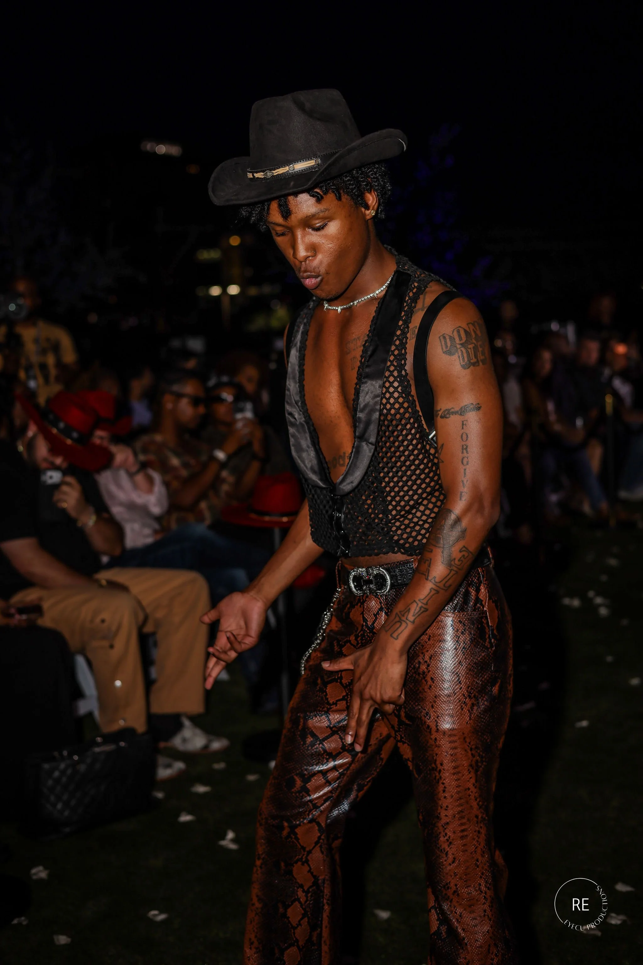 A person with dark skin wearing a black hat, lace top, and snakeskin patterned pants at night, surrounded by seated audience members.