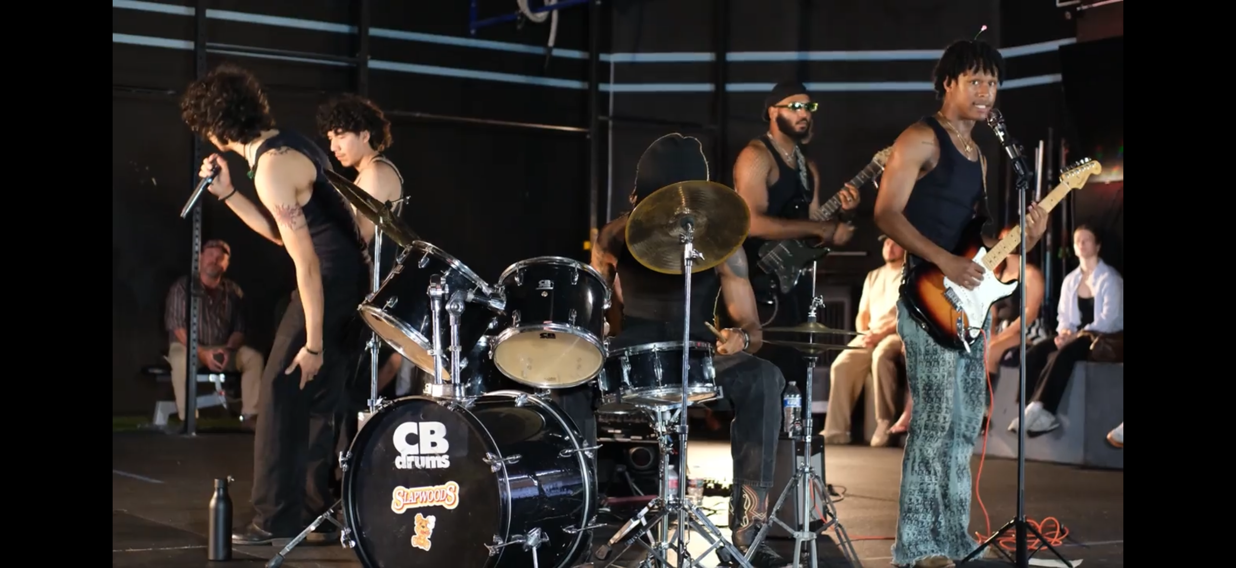 A live performance by a band on stage, featuring vocalists and musicians playing guitar and drums, with audience members seated nearby.
