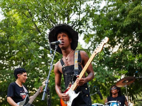 Musicians performing outdoors, with a woman singing into a microphone and playing an electric guitar, a man in the background playing a bass, and another person on drums, all in a wooded area.