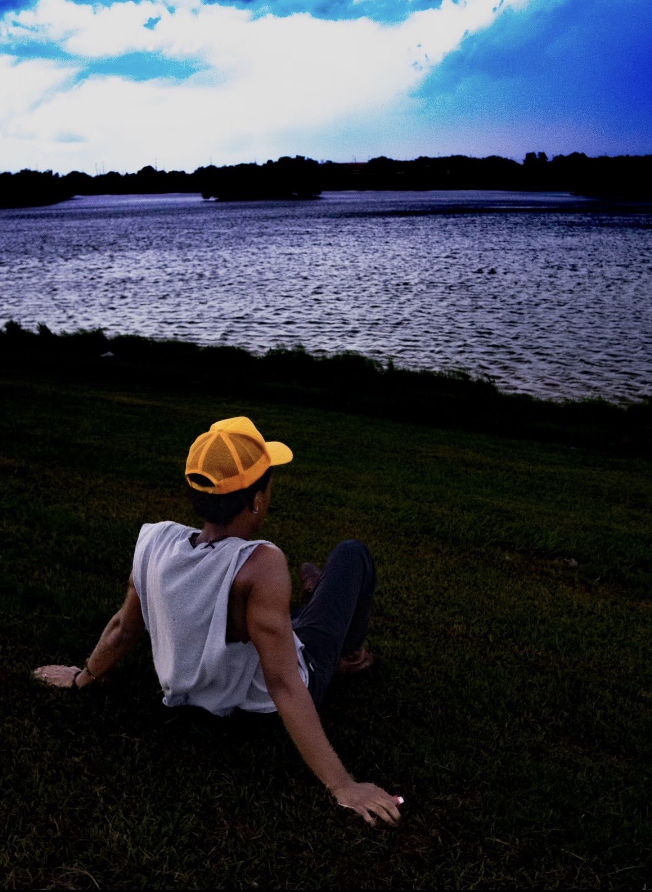 A person in a white shirt and yellow cap sitting on the grass near a large body of water, with trees and a cloudy sky in the background during dusk.