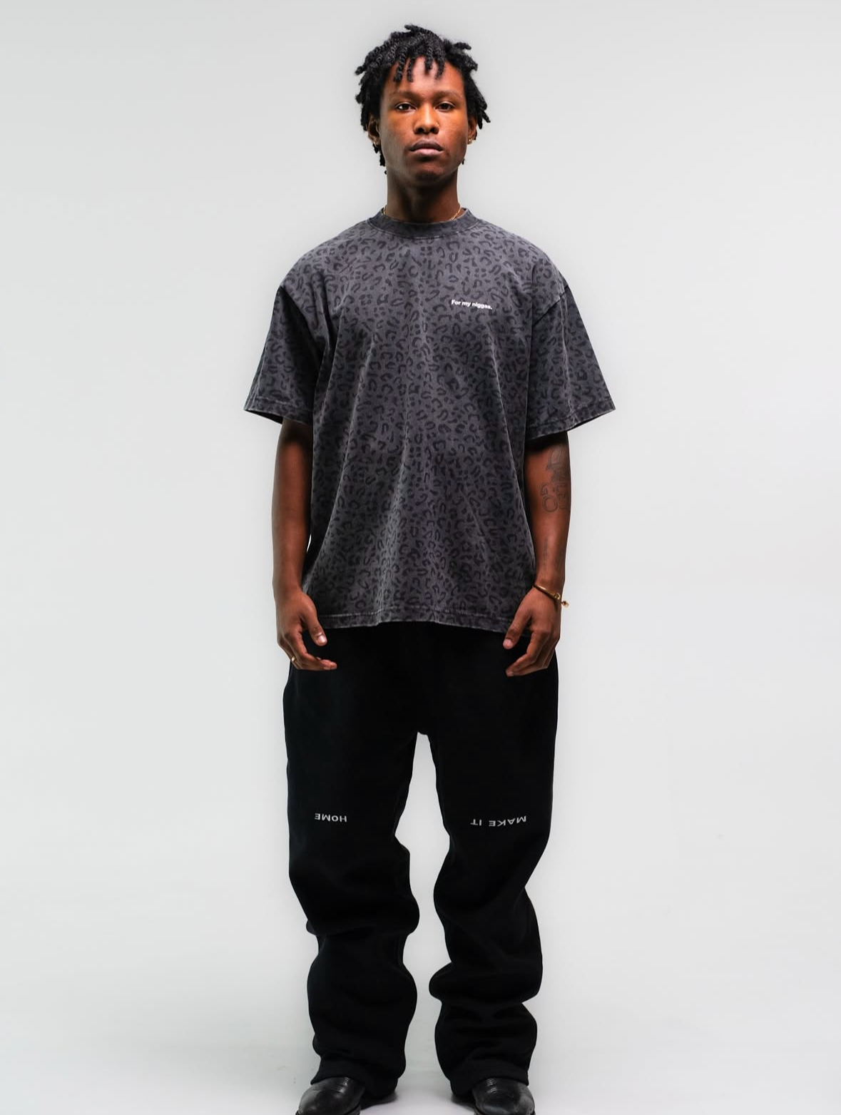 A man with dreadlocks stands against a plain light gray background, wearing a dark leopard-print t-shirt with white text that reads 'for my nipples' and black pants with white text reading 'MAKE IT HOME' on the left leg.